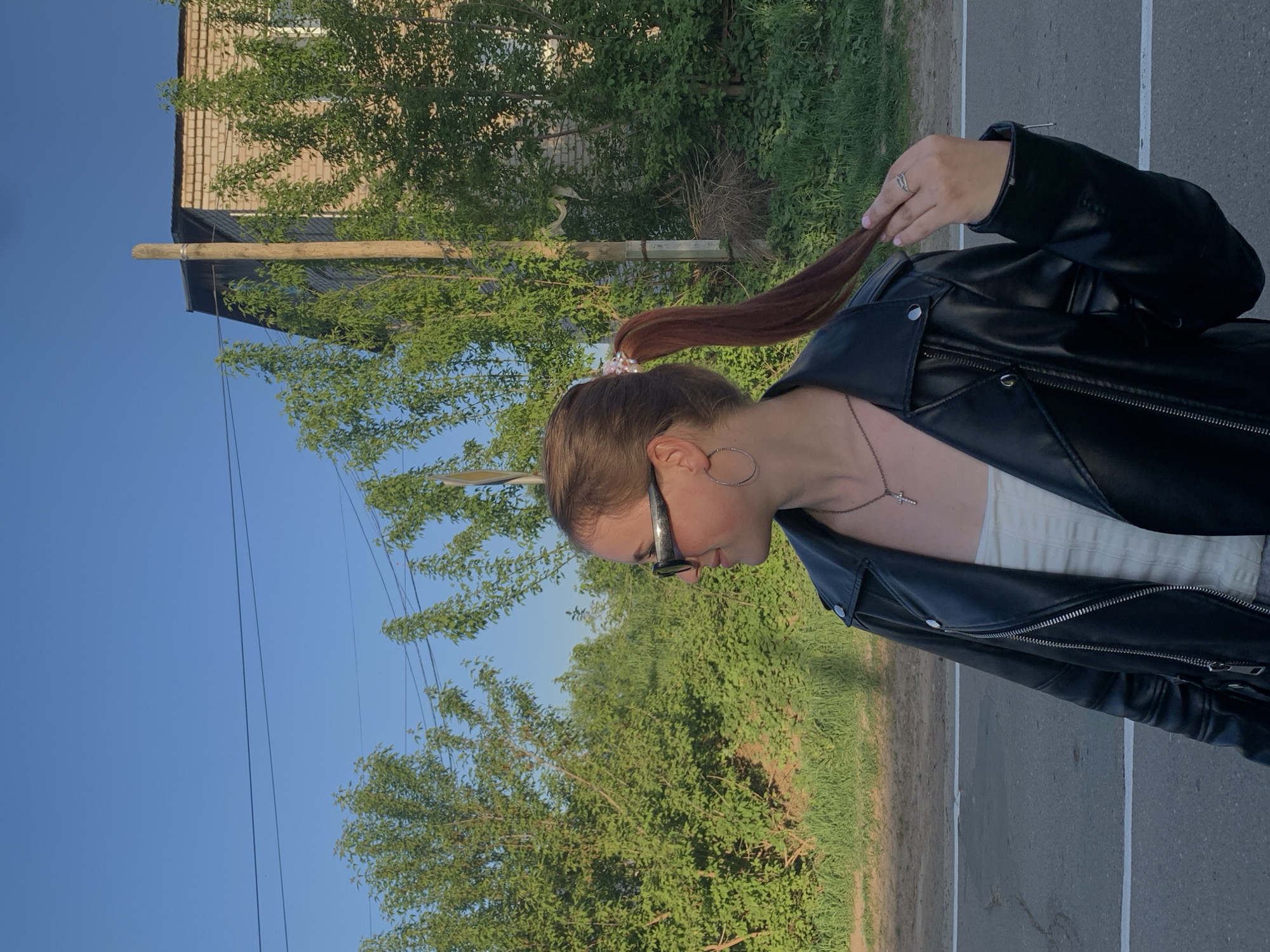 A medium shot depicts a woman standing outdoors in Фоки, Russia. She has brown hair pulled back into a ponytail, secured with a decorative hair tie. She is wearing a black leather jacket over a light-colored top, eyeglasses, and hoop earrings. A silver necklace with a cross pendant is visible. She is holding the end of her ponytail with her left hand, which has a ring on one of the fingers.
The background includes greenery, trees, a building, a utility pole, and a clear blue sky. The ground appears to be asphalt.