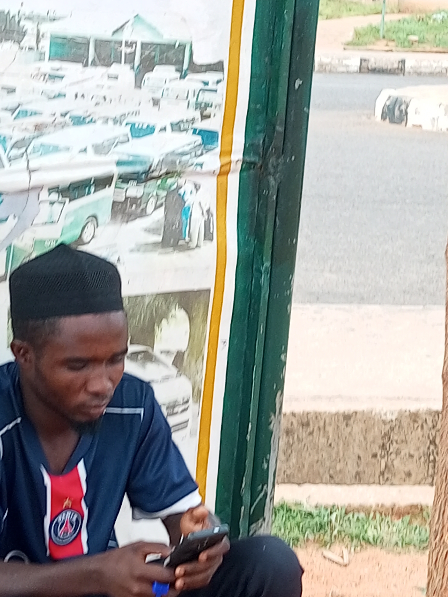 A young Black man wearing a blue and red jersey and a black kufi cap is seated outdoors, looking at his smartphone. He is positioned in front of a tall, green structure with yellow and white stripes, which appears to be a bus stop or shelter. Behind him, a poster displays images of multiple vehicles, including vans and cars, and some figures in traditional attire. The ground beneath him is dirt and some dry grass. In the background, a paved road is visible with white and gray concrete barriers, and beyond that, a dry, grassy area with some bushes. The overall setting suggests an urban or semi-urban environment, consistent with locations in Gombe, Nigeria.