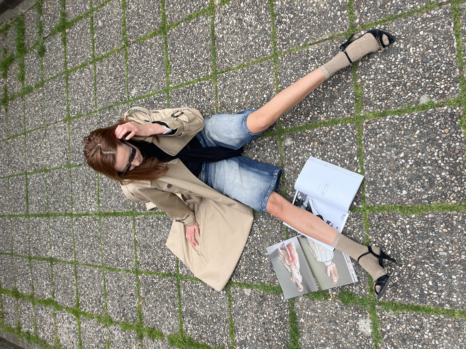 The scene is set on a paved surface within the city of Budva, Montenegro. An adult female is seated directly on a ground composed of square, grey, aggregate-textured concrete pavers with green grass growing in the seams. She is positioned with her legs extended forward in a wide V-shape. Her head is tilted downwards, and she appears to be looking at two open magazines or books placed on the ground in front of her.
The female is wearing a light tan trench coat, unbuttoned, over a black garment and a visible white shirt cuff. She wears faded blue denim jorts. On her lower legs, she has light beige ribbed socks, and over these, black open-toe high-heeled sandals. The left sandal features a gold-colored chain detail across the front strap. She is wearing dark-framed sunglasses. Her right hand is raised near her head, touching her hair or sunglasses. The two magazines on the ground show photographic content, with one displaying abstract or artistic imagery involving flesh tones and stripes, and the other displaying legible text on a light page. The lighting is soft and even.