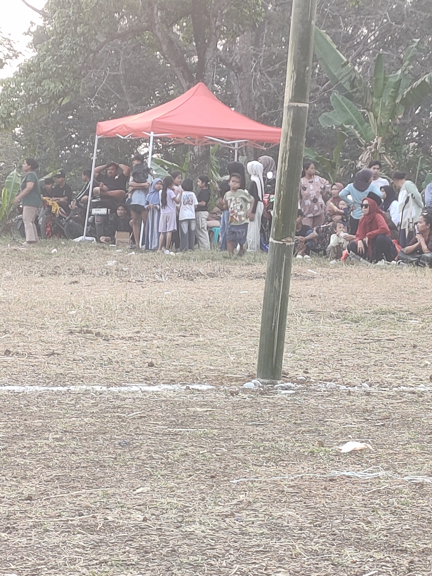 An outdoor gathering is taking place in Subang, Indonesia, near a field with dry grass. A red tent provides shade for a group of people, including adults and children, who are standing and sitting. A large bamboo pole stands vertically in the foreground. The people are dressed casually, with some children wearing bright clothing. The background shows trees and foliage, suggesting a rural or park-like setting. The overall atmosphere appears relaxed, possibly during a community event or festival.