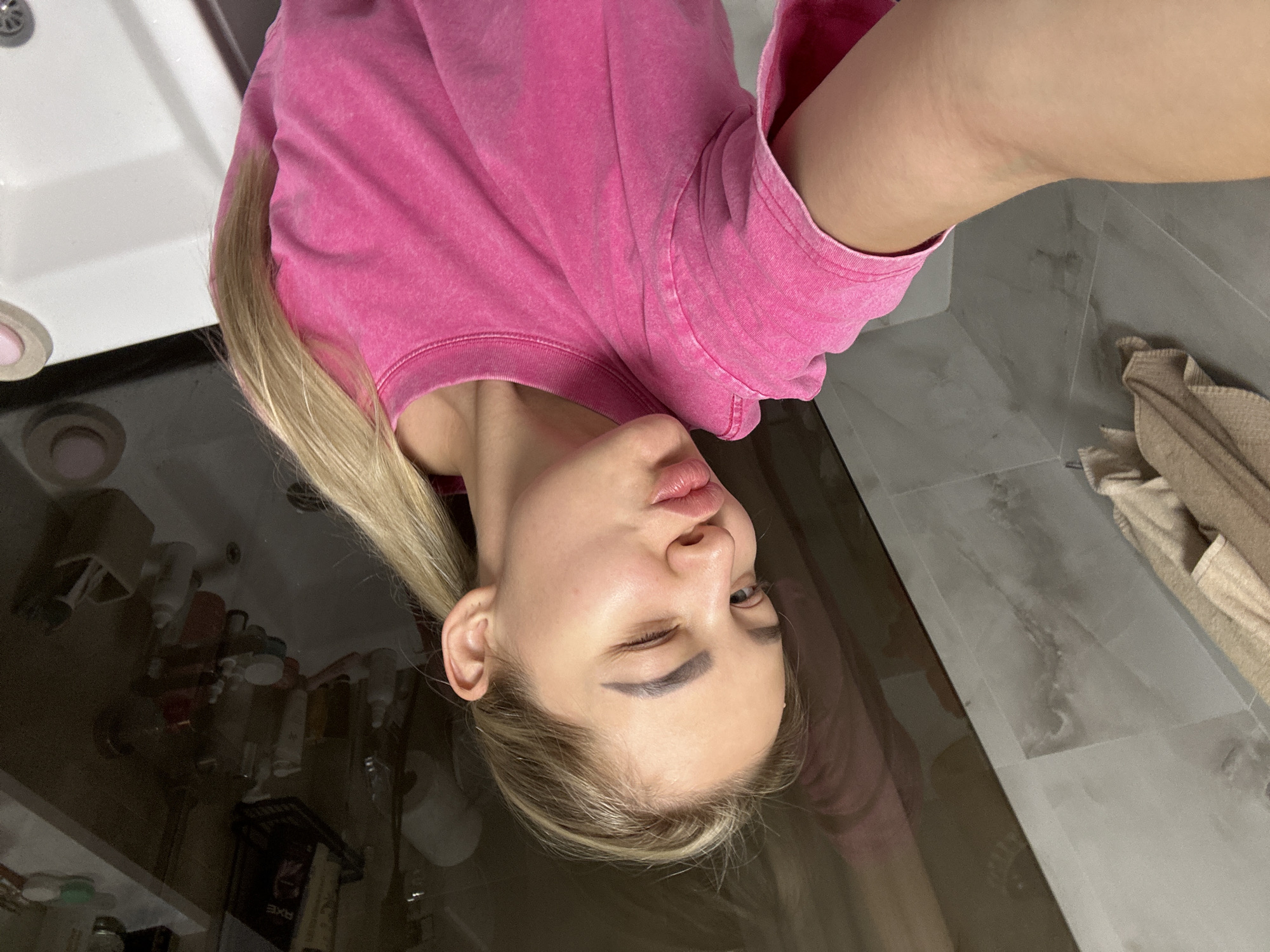 A young woman is positioned in a bathroom, possibly in a hotel room or apartment in Budva, Montenegro. The image shows her from a close, slightly angled perspective, suggesting a selfie taken by the individual. She's wearing a pink sweatshirt. The sink, floor, various bottles, and a towel are visible. The location inference comes from the general style of bathrooms in coastal apartments and hotels in Budva.