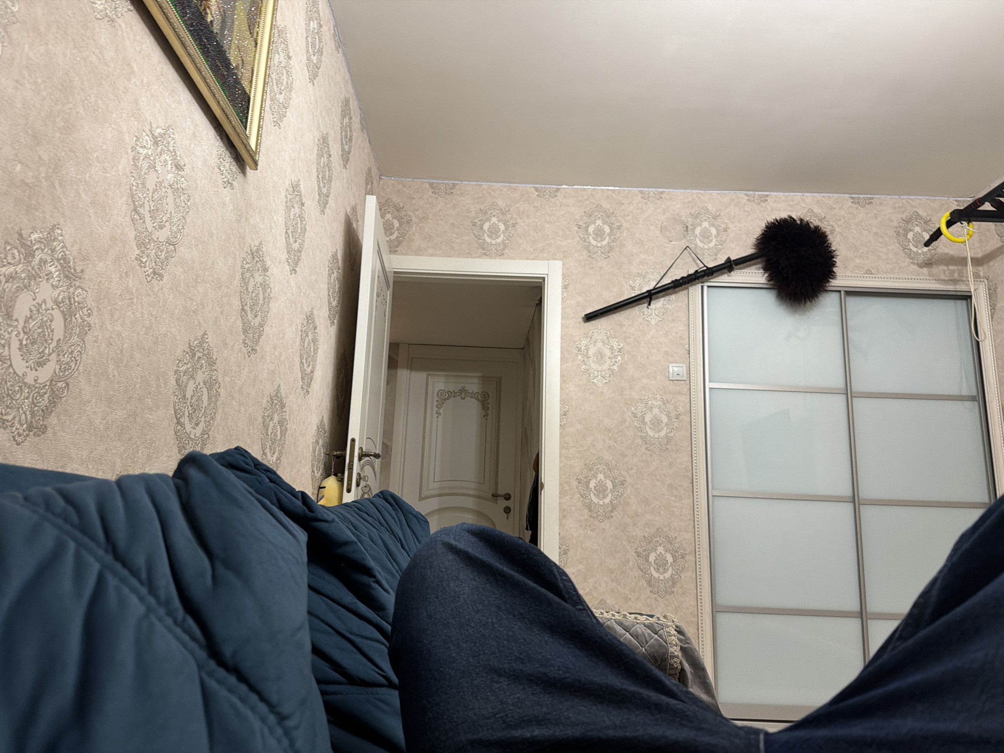 The image depicts an indoor scene from a first-person perspective, suggesting the viewer is seated or lying down. In the foreground, a portion of a person's dark blue jeans and the armrest of a dark teal-colored couch are visible. The couch is quilted with diagonal stitching. The background reveals a room with patterned wallpaper, a white door ajar, and a large closet with sliding doors. On the wall above the closet, a microphone boom with a windjammer is mounted, suggesting a filming or recording setup. A framed picture hangs on the wall to the left. The environment appears to be a residential interior. The location is not specified in the image, so the city is Unknown, Russia.