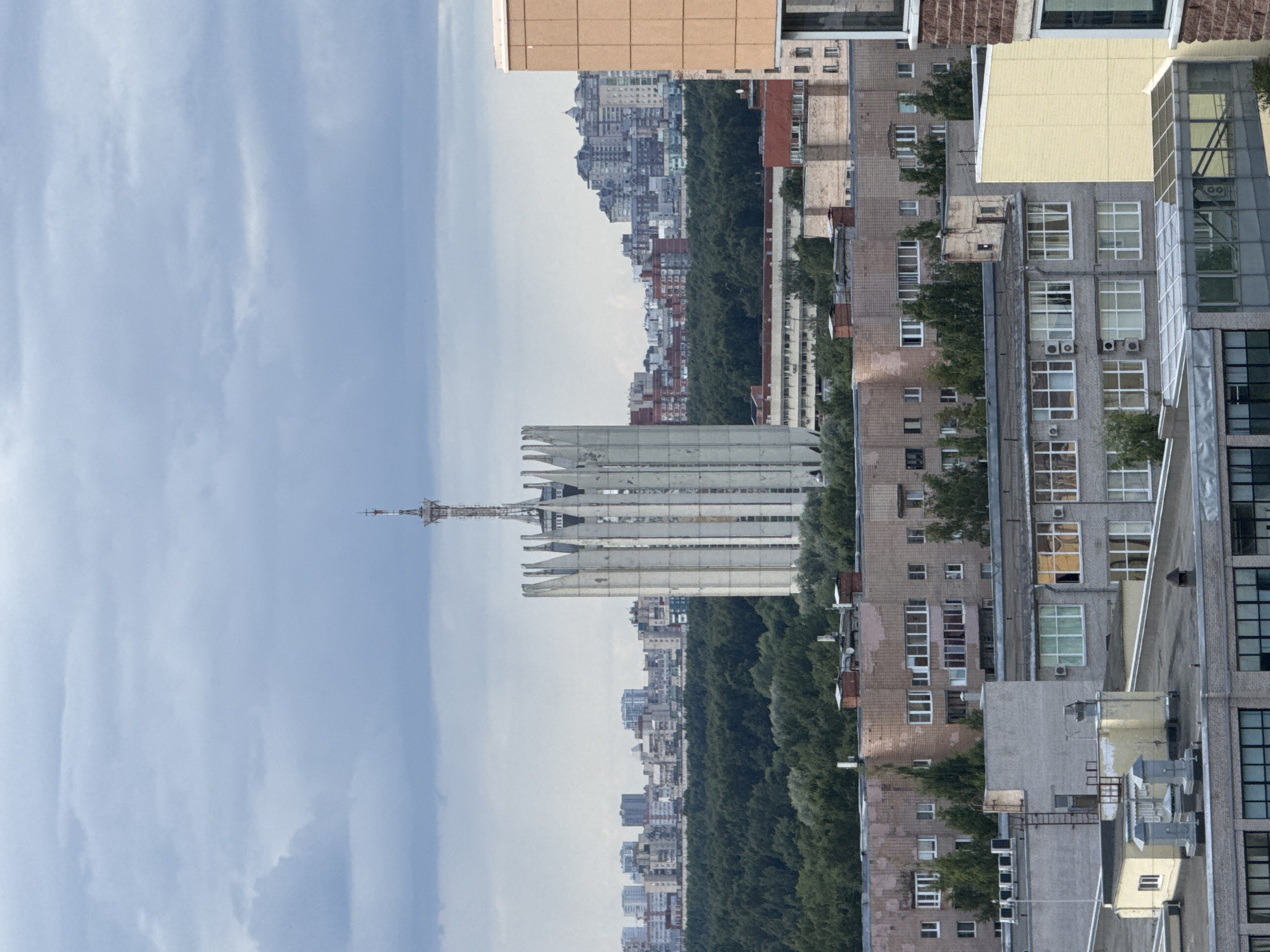The image shows a cityscape with a prominent, tall, white, angular tower in the center. The tower is topped with antennas. In the foreground, there are several multi-story apartment buildings and other structures. Behind the tower, a dense green forest separates it from a distant skyline of numerous apartment buildings of varying heights and architectural styles. The sky is overcast with a cloudy, muted gray appearance. The overall scene depicts an urban environment with significant green spaces and a notable central structure.