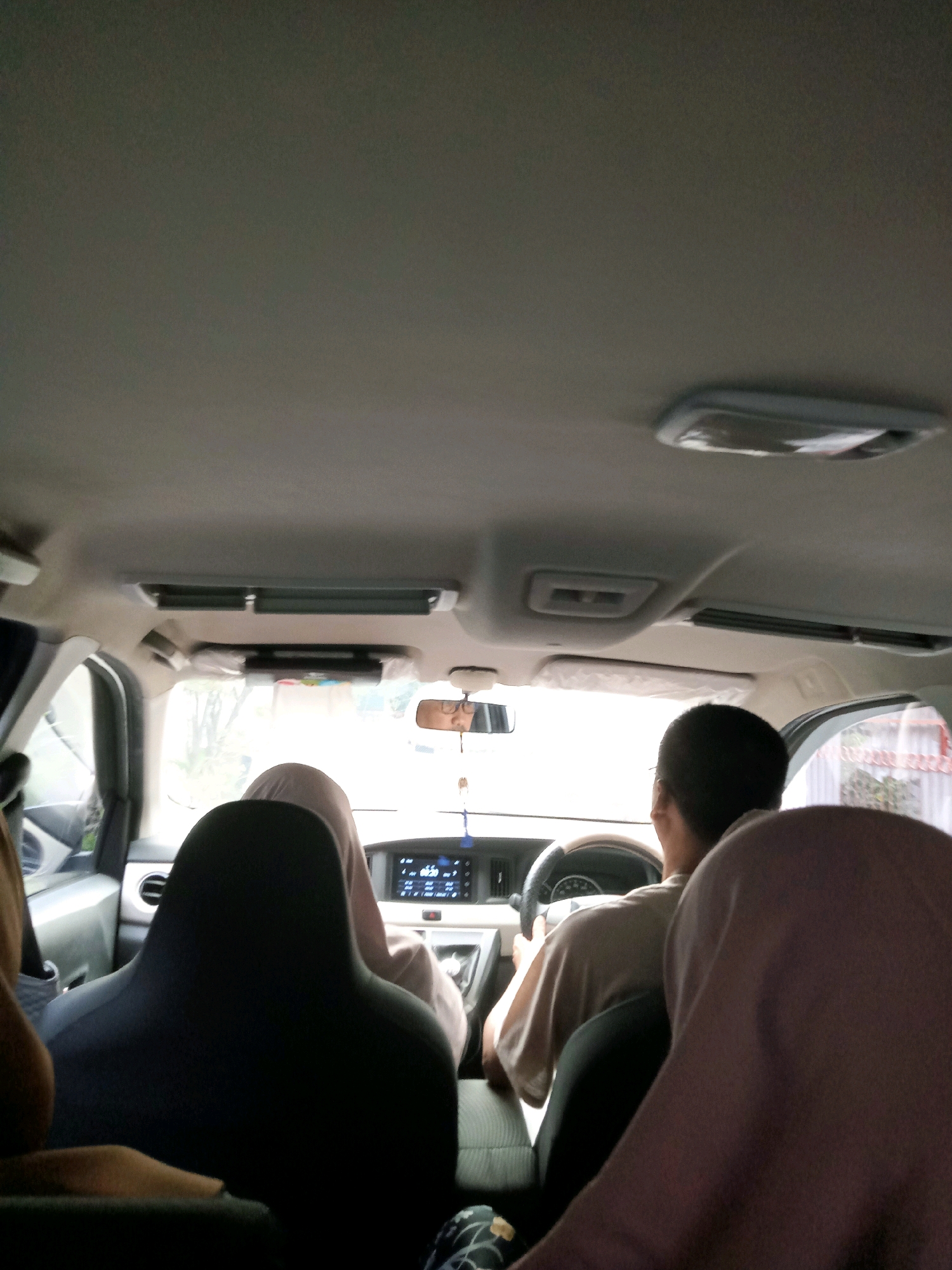 The image is taken from inside a car, looking forward. Three people are visible. A driver, a male with short dark hair, is in the front seat, hands on the steering wheel. To his left, a person wearing a beige hijab is in the passenger seat. In the back seat, the heads of two individuals are visible, one in a dark headrest and the other in a beige hijab. The car interior shows a dashboard with a screen, air vents, and a rearview mirror. Outside the car, trees and buildings can be seen, suggesting an urban environment. The lighting is natural, indicating it is daytime. The car is likely in Cirebon, Indonesia, based on the visual context.