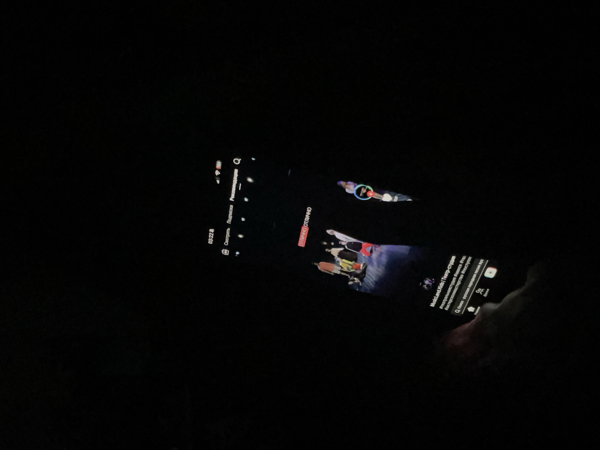The image shows a person holding a smartphone in a dark environment, possibly at night. The phone screen displays a video player interface, likely from a platform like YouTube, with Russian text. The video appears to be a performance or a rehearsal by a theater studio called "MusicLand.Kids" with hashtags related to acting and theater. There's also a search bar labeled "Q Поиск" (Q Search) and options for "Подписки" (Subscriptions) and "Рекомендации" (Recommendations). The scene suggests the person is engaging with digital content in a dimly lit setting, potentially at home or outdoors in a residential area given the location context of ЖК «Борисоглебское», Russia. The time displayed on the phone is 03:22.
