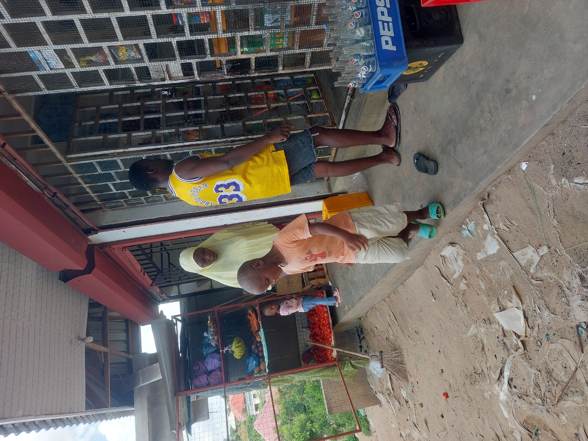 The image shows an outdoor scene in Dar es-Salaam, Tanzania. A young man in a yellow basketball jersey and denim shorts stands near a storefront with a caged front. A woman in a light yellow headscarf stands with a young boy in a peach-colored shirt. Behind them is a caged display of fruits and vegetables. A small child wearing pink is visible. A broom leans against the display. Cases of Pepsi bottles are stacked next to the storefront. The ground is dirt and littered with debris.