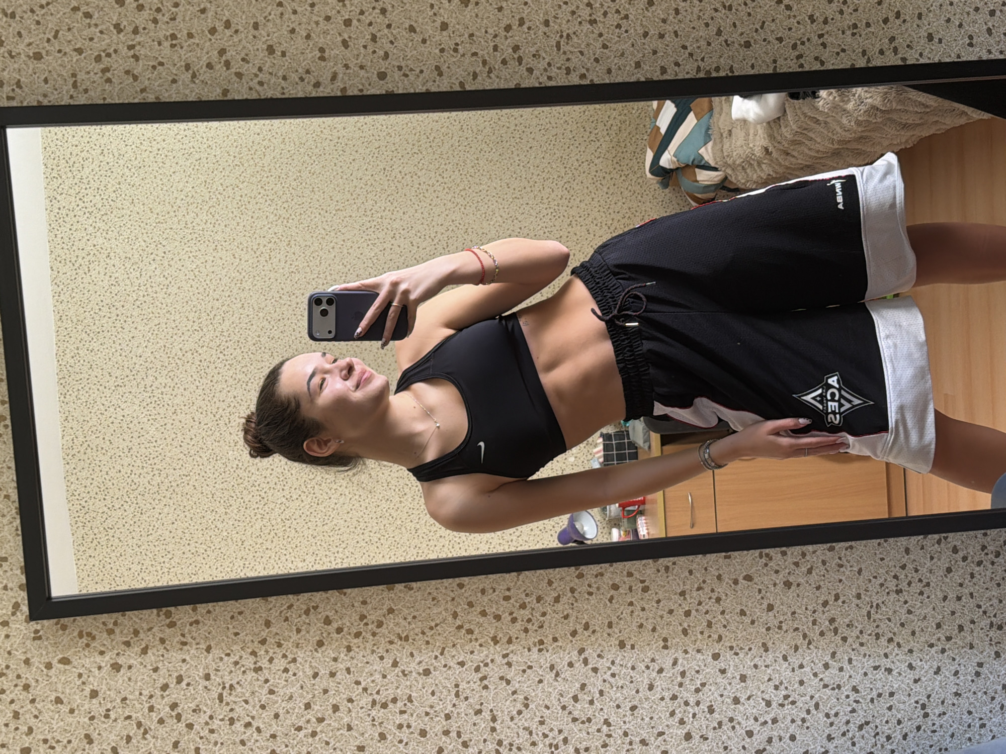 A woman is taking a selfie in front of a mirror. She is wearing a black sports bra and black athletic shorts. Her hair is pulled back in a bun. The mirror is hanging on a wall with patterned wallpaper. A cabinet is visible in the mirror, and items are on top of it. The location of the image is not specified; therefore, the city of Sofia, Bulgaria is not identifiable.