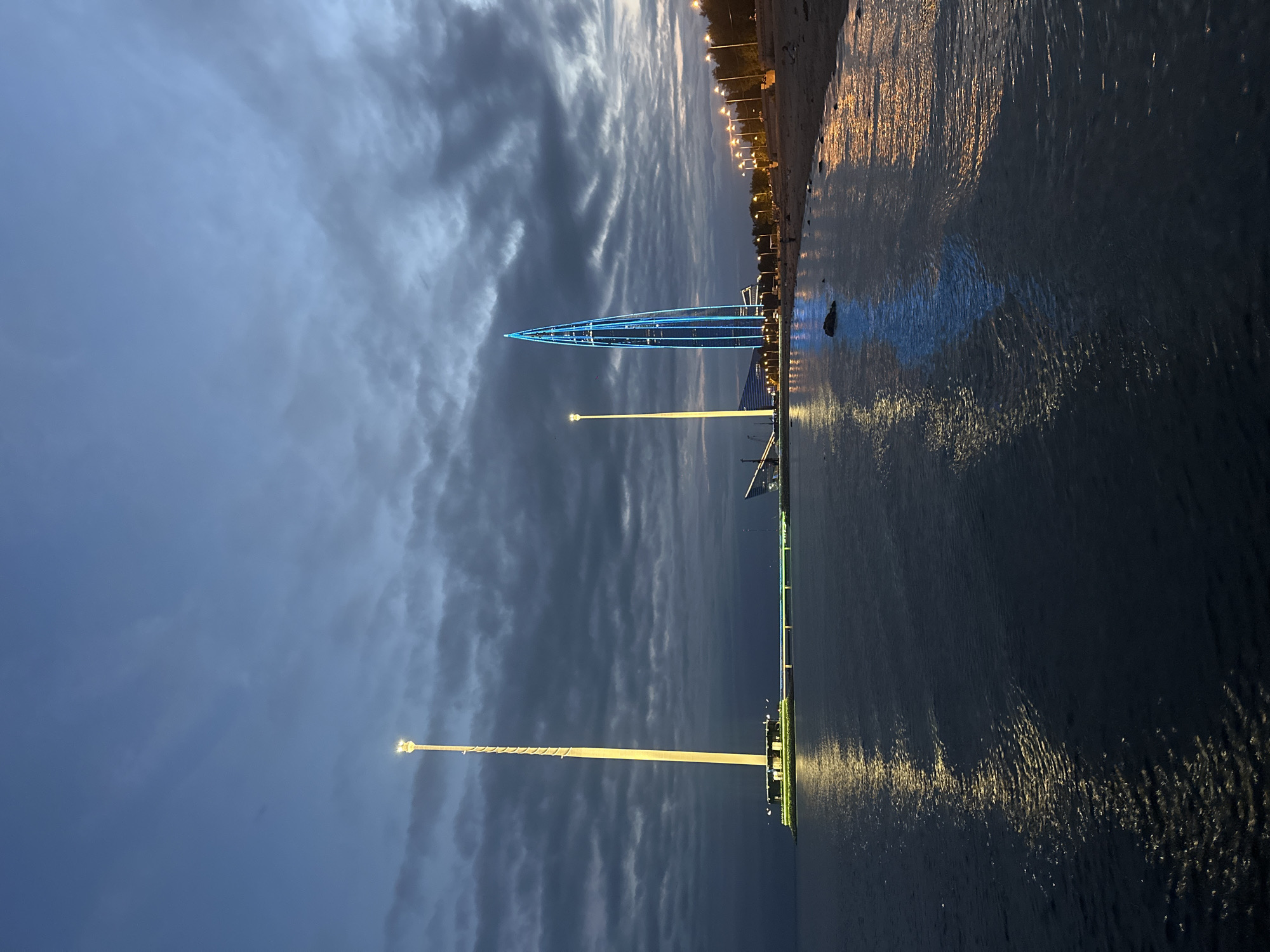 The image depicts the St. Petersburg skyline at dusk, featuring the Lakhta Center, a prominent skyscraper, illuminated with blue lights. To the left of the Lakhta Center, a tall, slender tower with yellow lights stands on a pier extending into the water. The foreground is dominated by a body of water, likely the Gulf of Finland, reflecting the lights from the shore and the structures. A beach with scattered rocks and vegetation lines the right side of the frame, with a row of streetlights visible in the distance. The sky is overcast with dark, dramatic clouds. No people are clearly visible in the image.