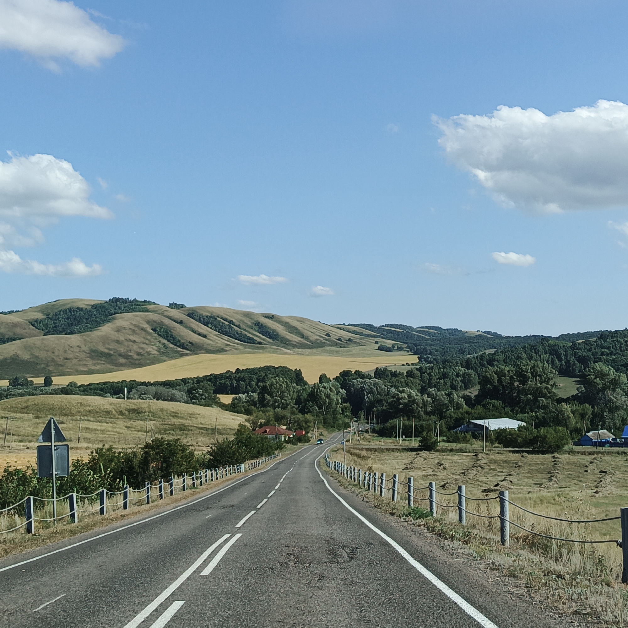 The image shows a paved road with white lane markings, stretching into the distance. The road is bordered by a white rope fence with metal posts on both sides. To the left of the road, there is a blue traffic sign on a post, and beyond that, a grassy hillside dotted with some trees. To the right of the road, there is a field with cut grass or hay, some scattered trees, and a few buildings including a white-roofed structure and a small blue building in the distance. Beyond the fields, rolling hills covered in greenery and yellow fields extend to the horizon. The sky is blue with scattered white clouds. The scene appears to be in a rural setting.