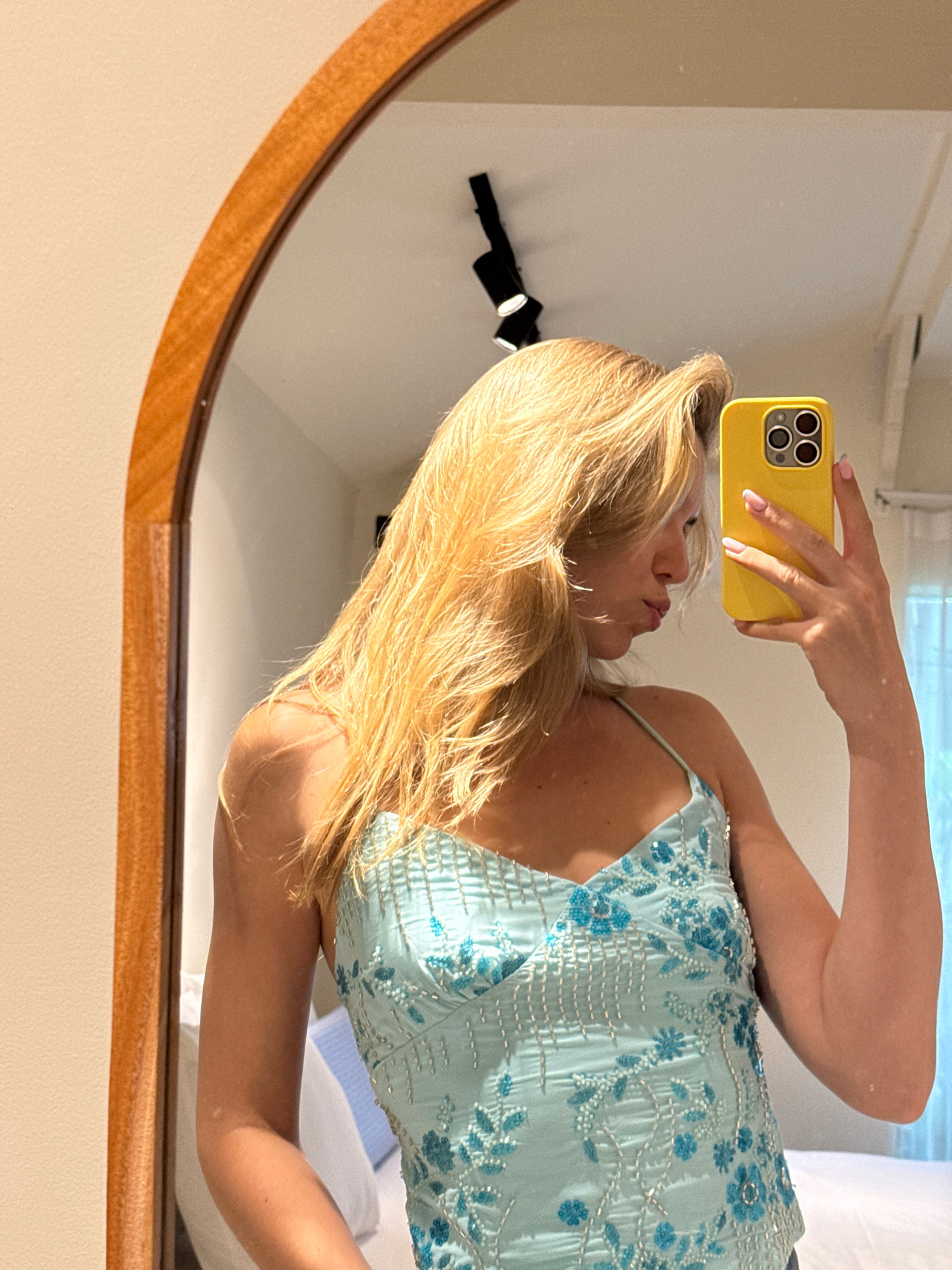 A blonde woman with long, wavy hair is taking a mirror selfie in a room. She is wearing a light blue tank top with blue floral embroidery and silver beads. She is holding a yellow iPhone in her right hand, and her lips are pursed. The mirror has a wooden frame, and a light fixture is visible on the ceiling in the reflection. The background is blurred, but a bed with white and blue pillows is partially visible.