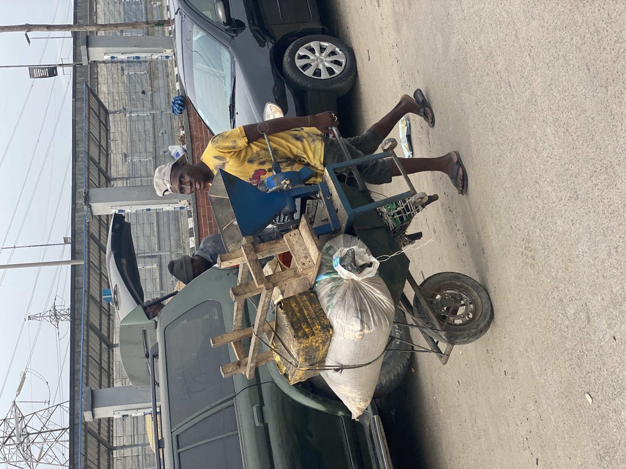 A man in a yellow shirt, grey shorts, and a light-colored cap is pushing a heavily laden handcart in Lagos, Nigeria. The cart contains a mix of items, including a metal grinding machine painted blue, wooden scaffolding, a yellow container, and large white sacks. The background features parked vehicles, including a dark-colored car and a green SUV. Overhead power lines and structures are visible in the background, indicating an urban setting. The ground is paved.