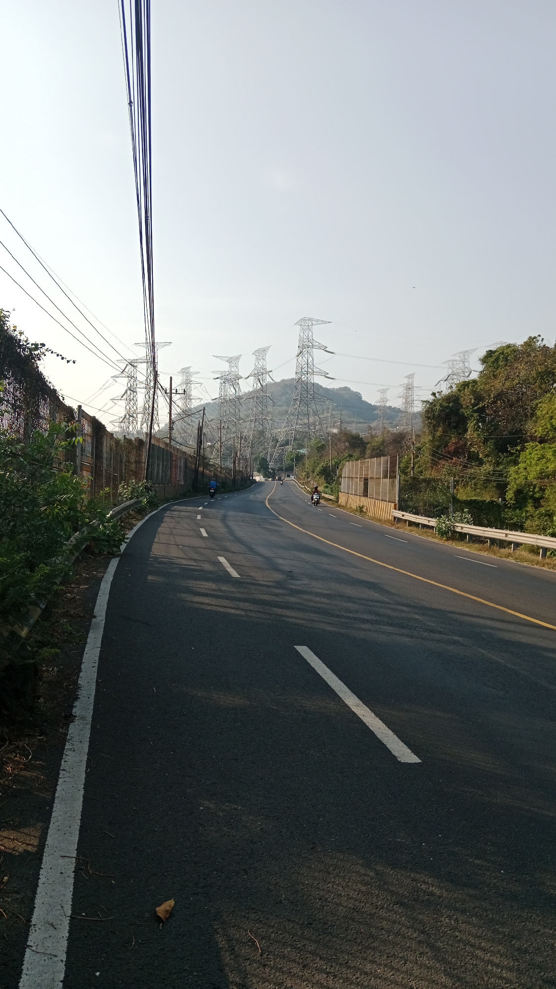 The image shows a view of a road in Bhinar, Indonesia, with numerous tall power line towers extending up a hillside in the background. The road is paved and has white lane markings, with a yellow line dividing lanes. Several motorcycles are visible on the road. On the left side of the road, there is a metal fence bordering lush green vegetation. On the right side, there is more vegetation and a sound barrier made of beige panels. The sky is clear and light, suggesting it is daytime. The power line towers are a prominent feature of the landscape.