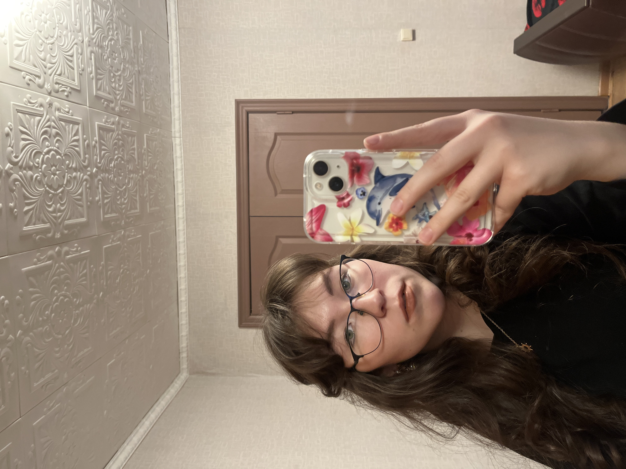 A young woman with long wavy brown hair is taking a selfie in what appears to be an indoor environment. She is wearing black-rimmed glasses and a black top, and a gold-colored necklace is visible. The background includes a decorative wall panel with an ornate floral pattern, a light-colored wall with a subtle pattern, and a brown wooden door or frame. A shelf-like object is visible in the upper right corner, with a red item on it. The woman is holding a smartphone with a floral-themed case in front of her face. The selfie is taken at an angle. There is no specific information that could identify the precise location as being in Izhevsk, Russia.