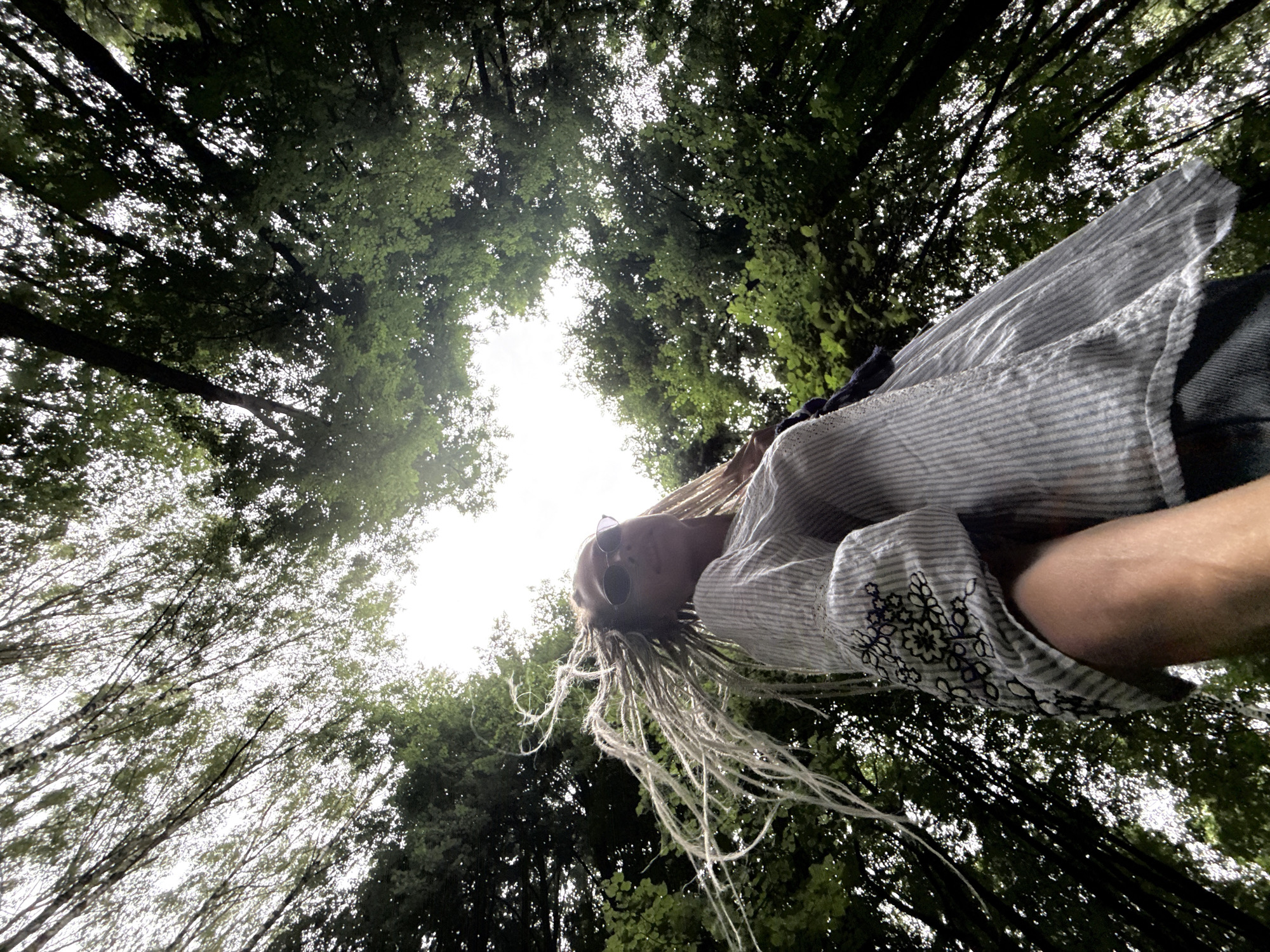 A low-angle shot looks up through the canopy of trees in Rusanovo, Russia. The leaves and branches create a dense, green ceiling against a bright, overcast sky. A person with light-colored dreadlocks and sunglasses is visible from the chest up, looking upwards and slightly to the right. They are wearing a striped shirt with floral embroidery on the sleeve. The person's hair is blowing in the wind, creating a dynamic element against the static backdrop of the trees. The scene appears to be in a natural, outdoor setting, possibly a park or forest.