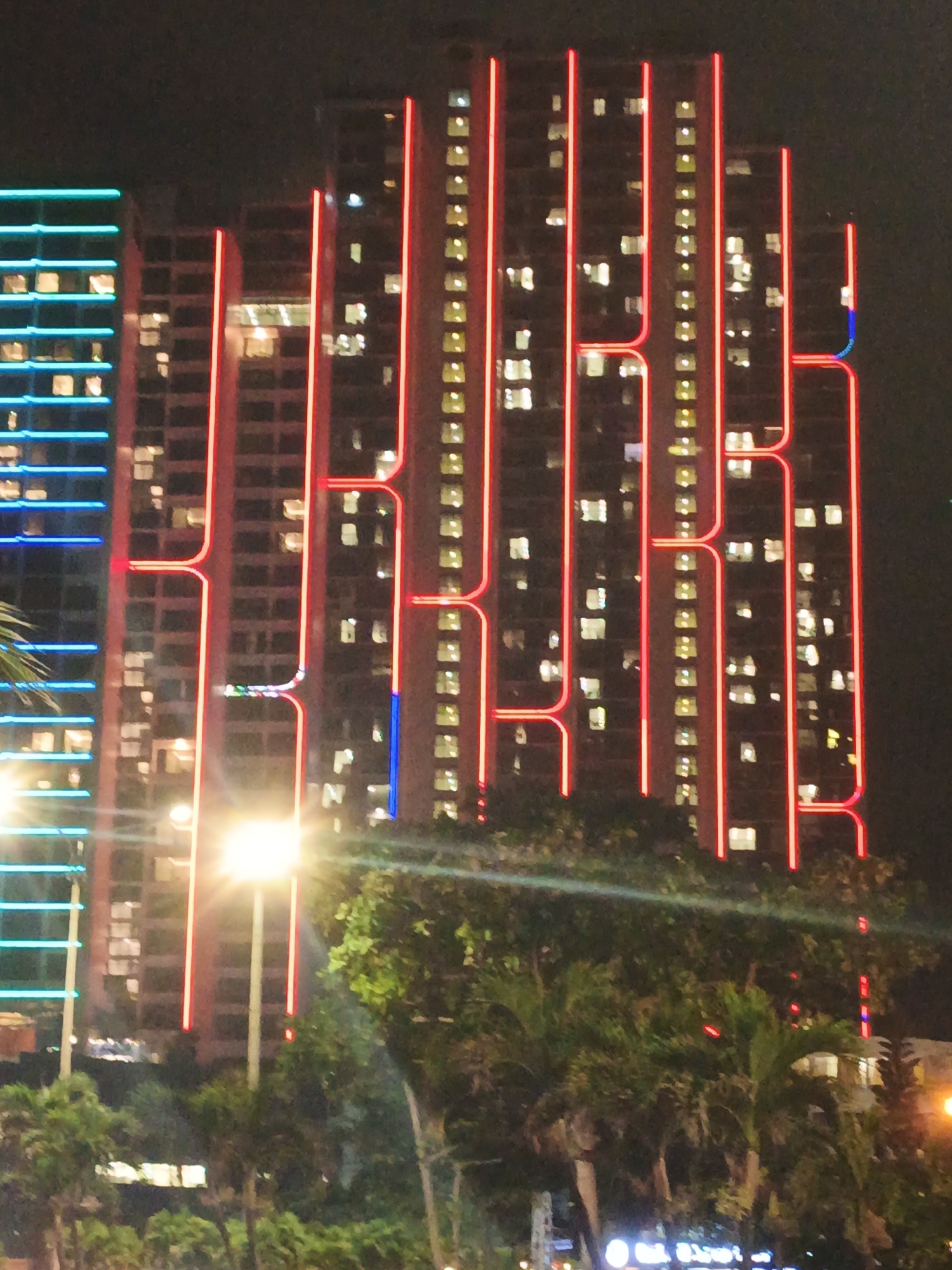The image captures a nighttime view of several high-rise buildings in Nha Trang Ward, Vietnam. The buildings are illuminated with bright neon lights, primarily in red and blue, outlining their structures and creating a visually striking urban landscape. The red lights form a distinctive pattern that suggests lettering or design elements on the facades of the buildings. One building on the left features horizontal blue lights. The foreground is dominated by lush green trees, some appearing to be palm trees, obscuring the lower portions of the buildings and the ground level. A bright streetlight is prominent in the center of the image, casting a strong glow. The overall scene depicts a vibrant and modern city at night, characterized by architectural lighting and tropical vegetation. There are no visible people or discernible actions taking place.