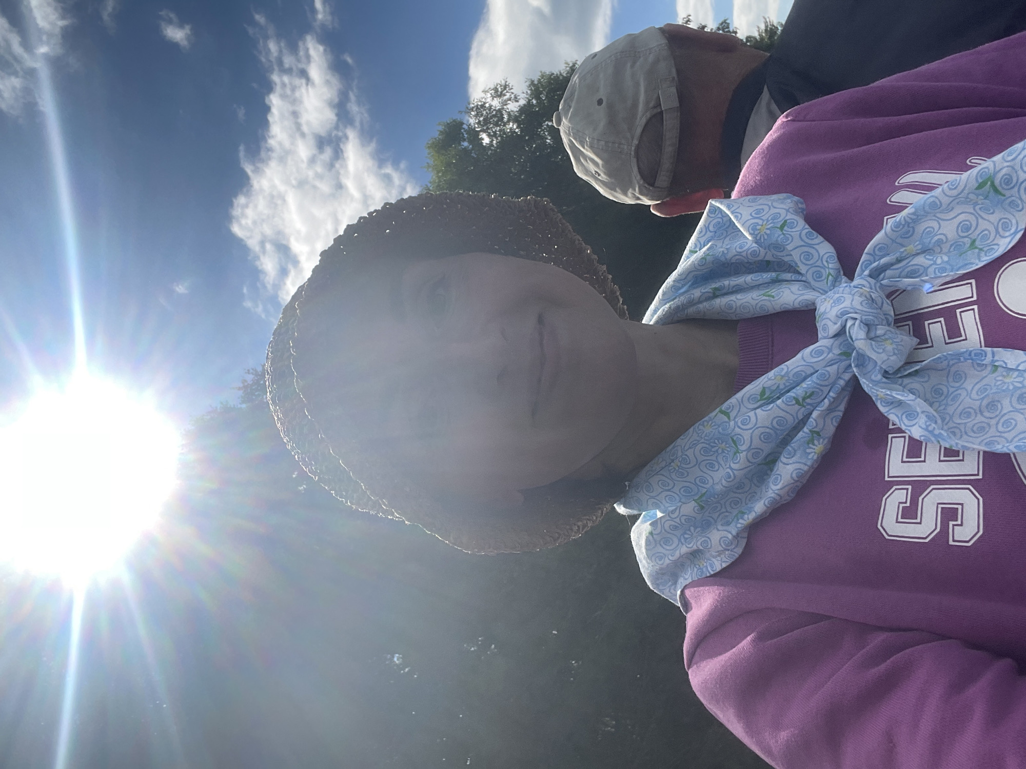 The image is a selfie taken outdoors under a bright sun. A woman wearing a straw hat, a purple sweatshirt, and a blue patterned scarf is in the foreground, looking towards the camera. Behind her, a man wearing a beige baseball cap and a dark jacket is visible from the back. The background shows trees and a blue sky with clouds. There is no information in the image that indicates the location is Dresvyanovo, Russia.