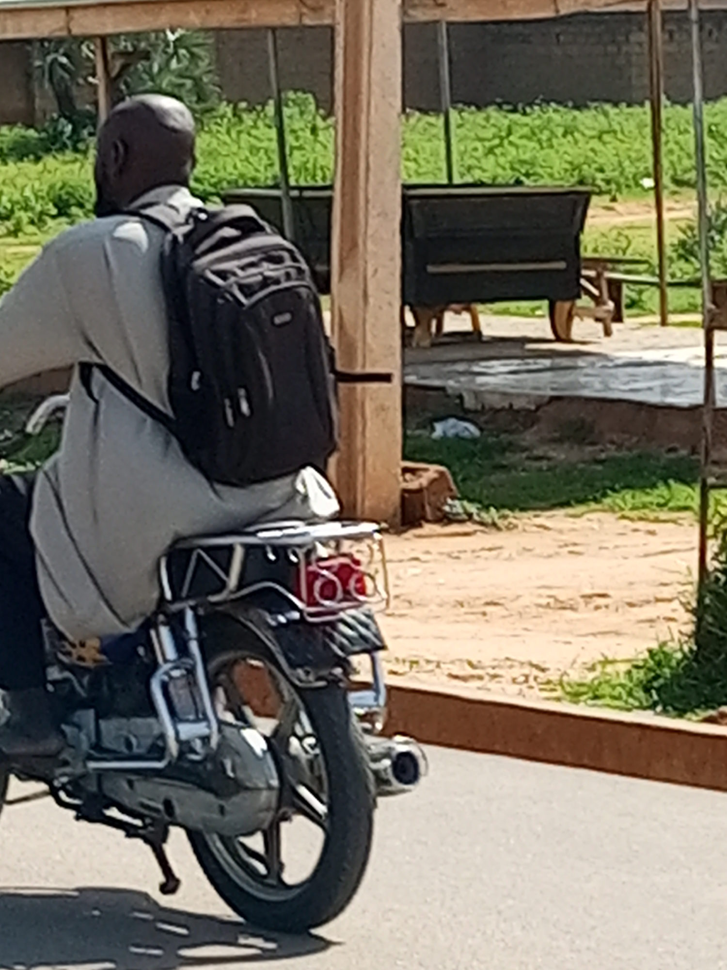 A man with dark skin and a shaved head is riding a motorcycle, carrying a black backpack. He is wearing a light-colored, long-sleeved garment. The motorcycle is black and silver with a visible engine and kickstand. Red taillights are visible on the rear of the motorcycle. The man appears to be in motion on a paved road. In the background, there is green vegetation, a dark-colored object with wheels resembling a cart or stall, and wooden structures. The environment suggests an outdoor, possibly semi-urban or rural setting.