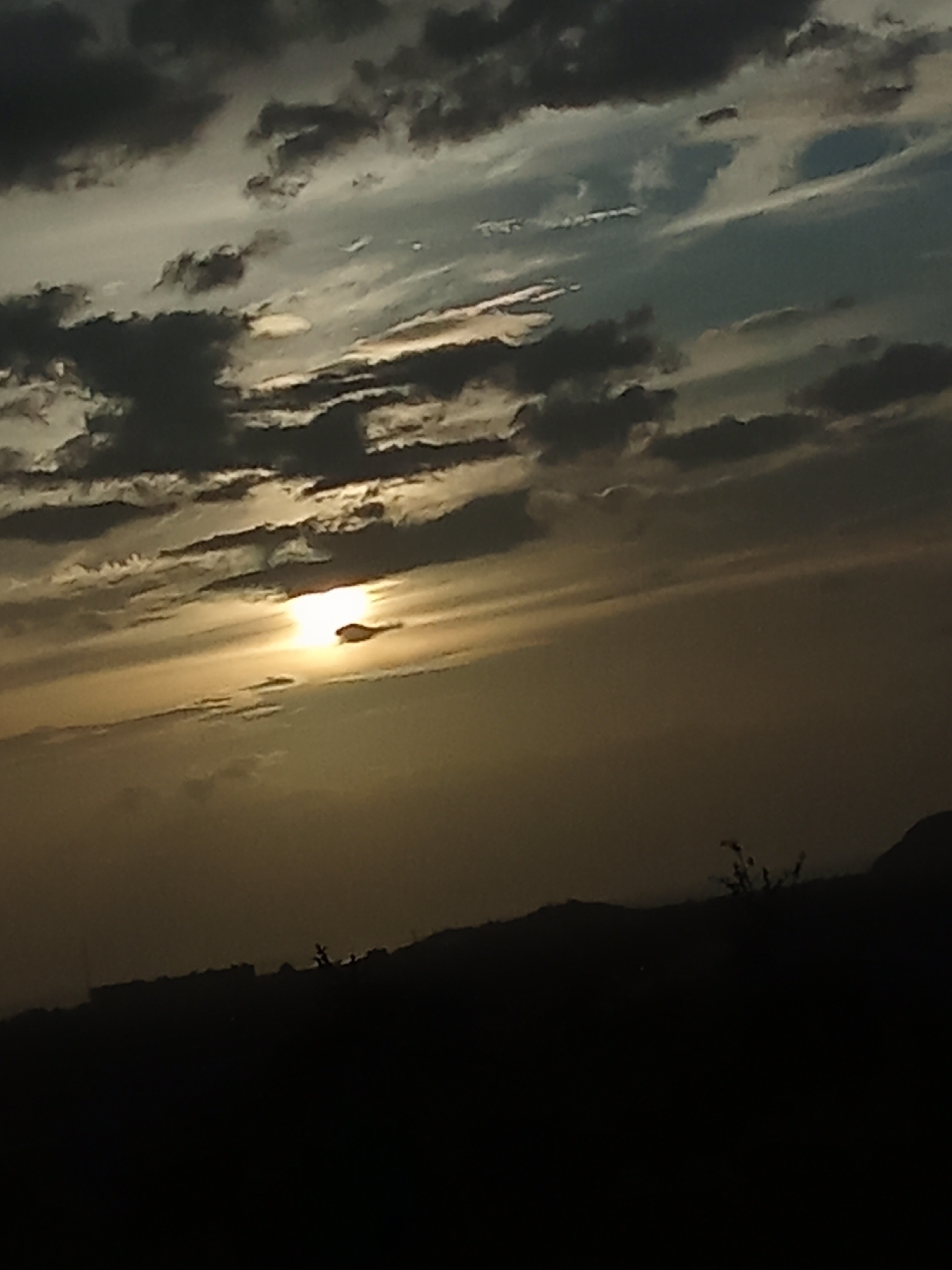 The image shows a landscape silhouette at sunset. A dark, indistinct hill line with some sparse vegetation and structures (possibly buildings) is visible at the bottom. Above, the sky is filled with layered clouds. The sun is partially obscured by clouds, creating a bright golden glow. This scene could be located in Juan Griego, Venezuela due to its coastal location and hilly terrain. There are no people visible in the image.