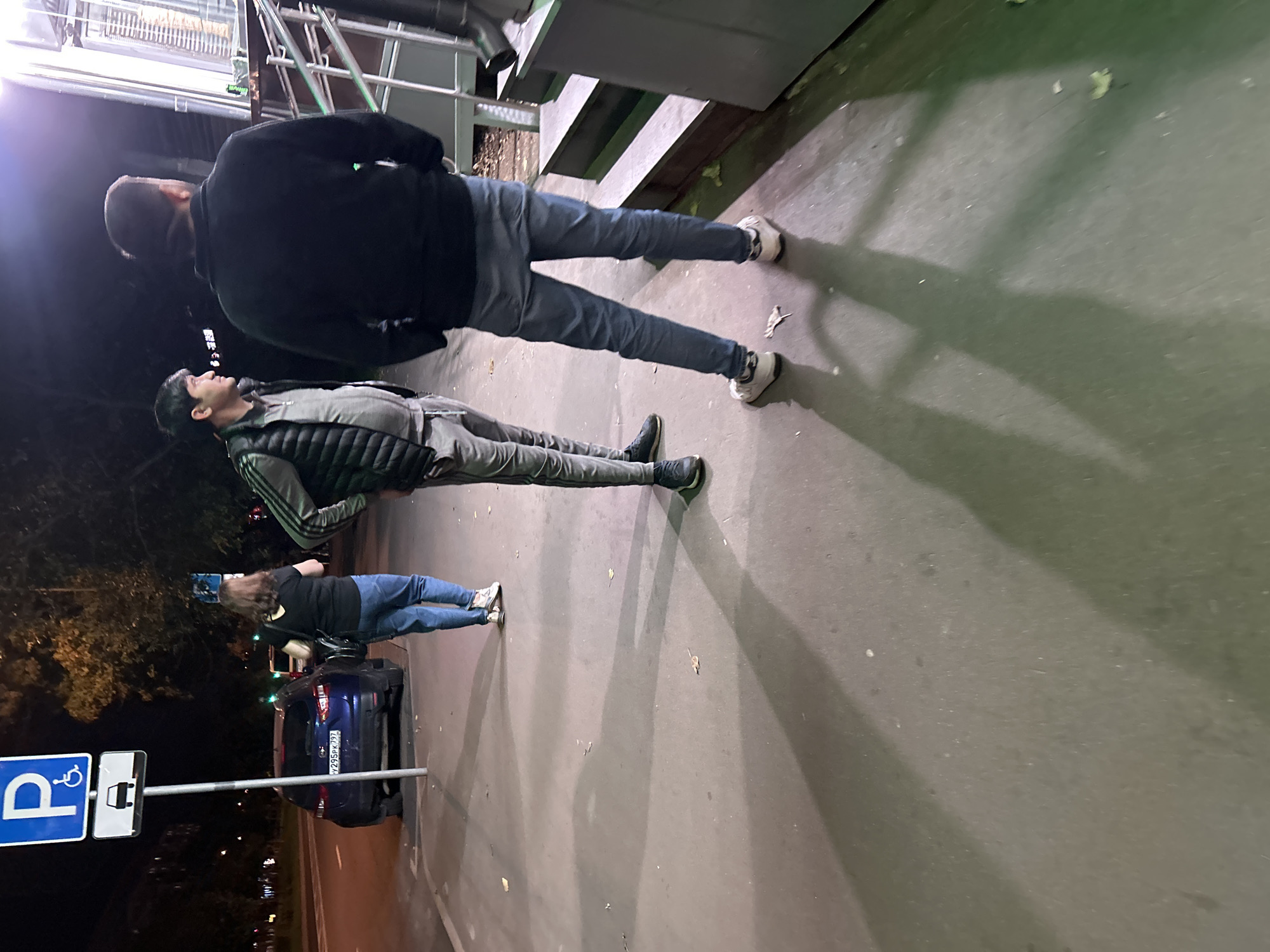The image depicts a street scene at night in Moscow, Russia. In the foreground, two men are standing on a sidewalk. The man on the left is wearing a grey tracksuit with a dark vest over it, and the man on the right is wearing a dark jacket and jeans. Behind them, a woman in jeans and a black top is walking away from the camera. She is carrying a bag. To the left of the woman is a blue car, and a parking sign with a disabled person symbol and a car symbol is visible next to the car. The background shows a dark street with some trees and building lights. The overall atmosphere is casual, with the subjects appearing to be in transit or waiting.