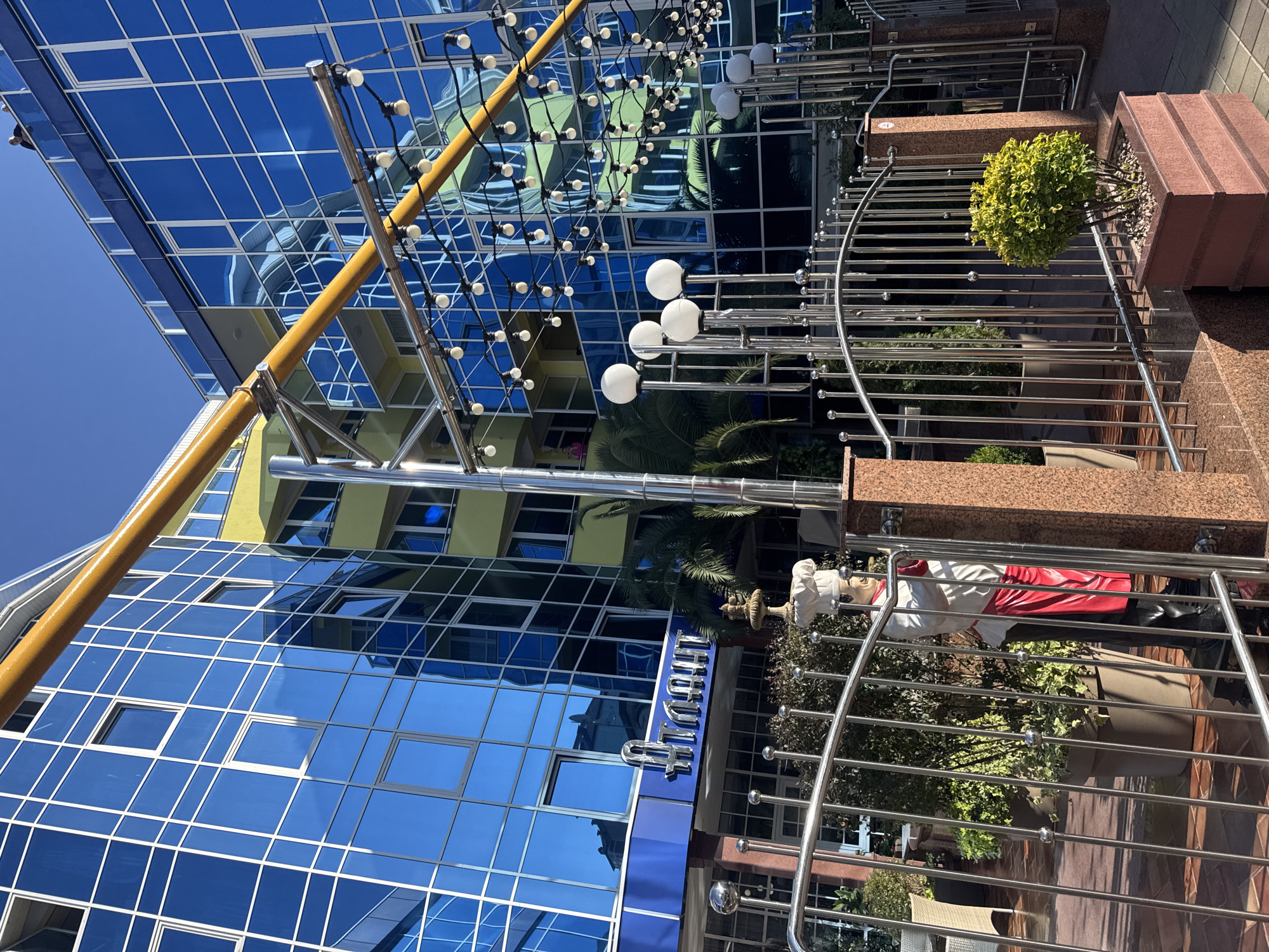 A chef statue stands behind a metallic fence in front of a modern building with blue reflective glass windows, located in Sochi, Russia. The building has a yellow section on its upper floors and is adorned with a string of lights. The scene is illuminated by sunlight, casting shadows on the ground.
