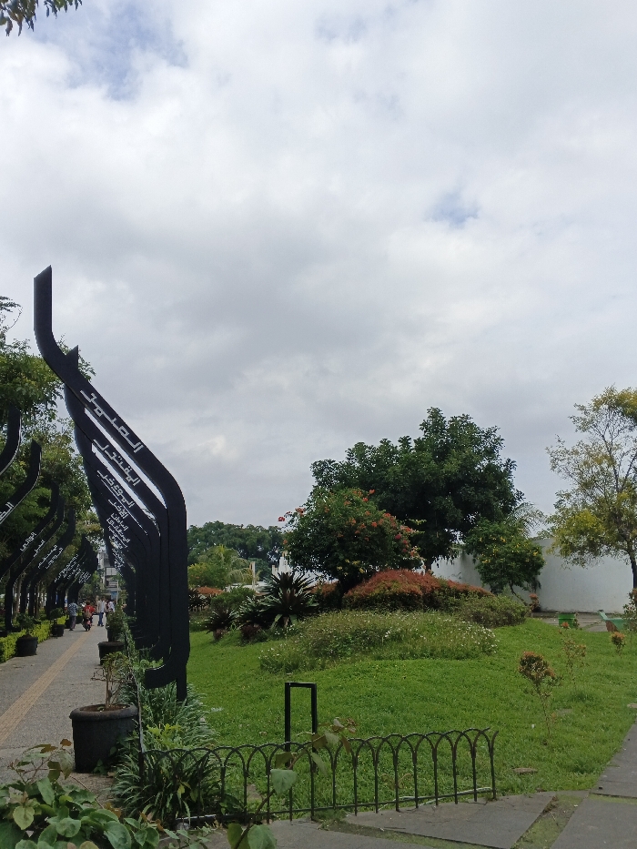 The image depicts a public park area in Bien Hoa, Vietnam, under an overcast sky with diffuse, light grey clouds. On the left, a paved pedestrian walkway extends into the background, lined by a series of tall, black, curved upright structures. Each structure features vertically oriented white text, identifiable as Arabic script, with words such as "الصلو", "القتلي", "البعض", and "اليوم" visible. Several dark potted plants are positioned along the walkway. In the mid-ground on the walkway, a small group of indistinct individuals is observed walking. Trees with green foliage form a canopy above and behind these structures. The right side of the image consists of a well-maintained green lawn. Various shrubs and trees are distributed across the lawn, including a prominent cluster with dense green and red foliage in the center-right. A low, dark green metal fence with arched segments is visible in the foreground, separating a small patch of ground from the main lawn. In the background on the right, a light-colored building or wall is partially obscured by trees.