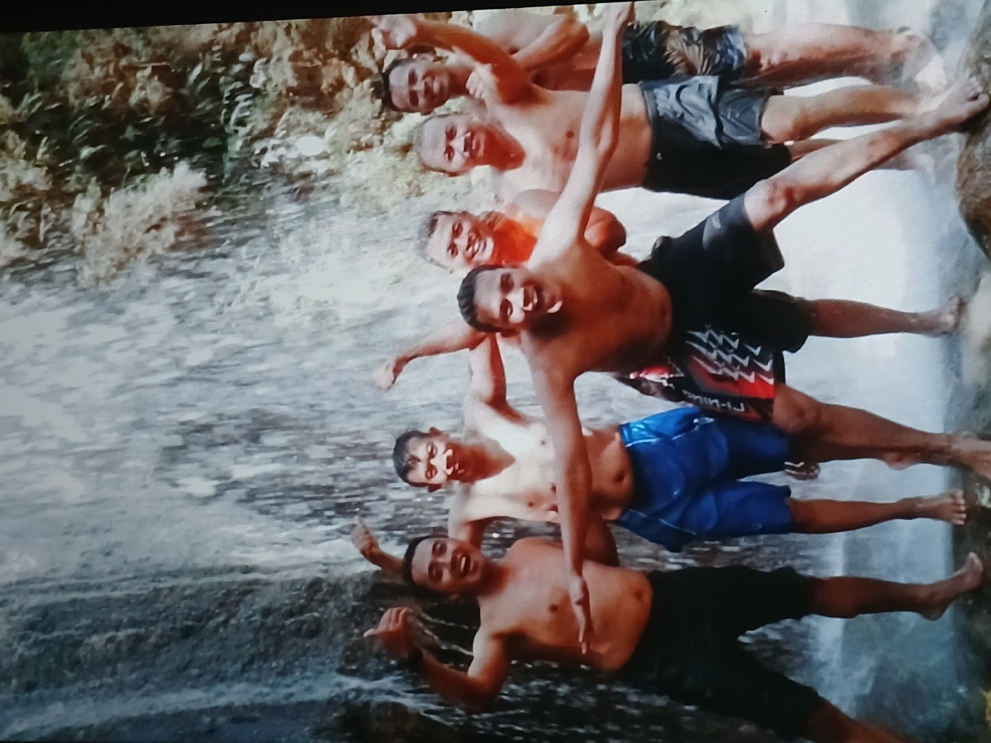 The image depicts a group of six shirtless men standing in front of a waterfall, some of them appearing to be in water. The men are wearing various swim shorts. The environment appears to be a natural setting with a waterfall cascading down a rocky and vegetated cliff face. The lighting suggests it is daytime. There is no visual information within the image to place this scene specifically within the city of Surabaya, Indonesia.