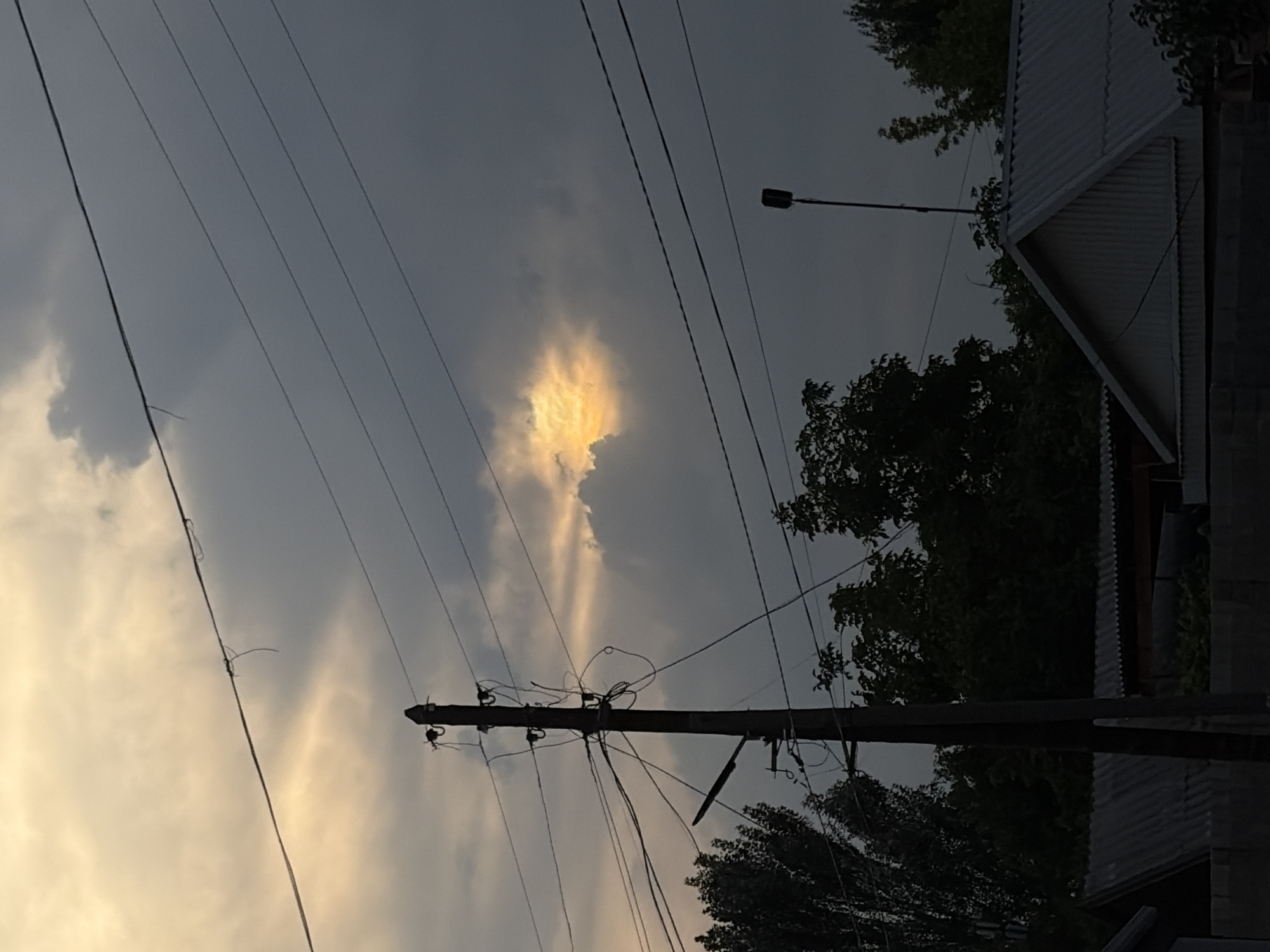 This image depicts a slightly overcast sky with dramatic cloud formations. A patch of bright, illuminated cloud is visible in the upper left quadrant, suggesting sunlight breaking through. Several power lines crisscross the frame, some appearing tangled. A utility pole with insulators and wiring stands prominently in the center, extending upwards. In the background, the corrugated metal roof of a building and dark green trees are visible on the right side. A street lamp is mounted on a pole in the upper right. The overall mood is somewhat somber due to the heavy clouds, but the illuminated cloud offers a touch of hope or drama. The scene appears to be set in a residential or semi-urban area, possibly in the late afternoon or early evening given the quality of light. There is no visible text in the image. The rotation of the image suggests it may have been captured vertically.