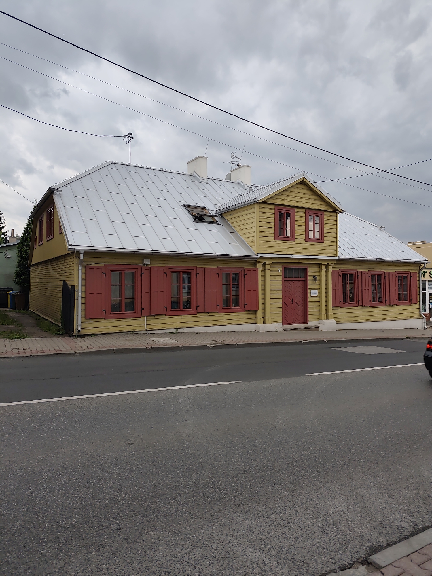 An exterior, eye-level, full shot captures a traditional, yellow wooden house with a silver, pitched roof in Zgierz, Poland. The house features red-shuttered windows and red double doors. To the left of the house, a dark green hedge and garbage bins are visible. The house is situated alongside a gray asphalt road, separated by a pedestrian sidewalk made of red and gray pavers. Overhead, a cloudy sky is crisscrossed by electrical wires.