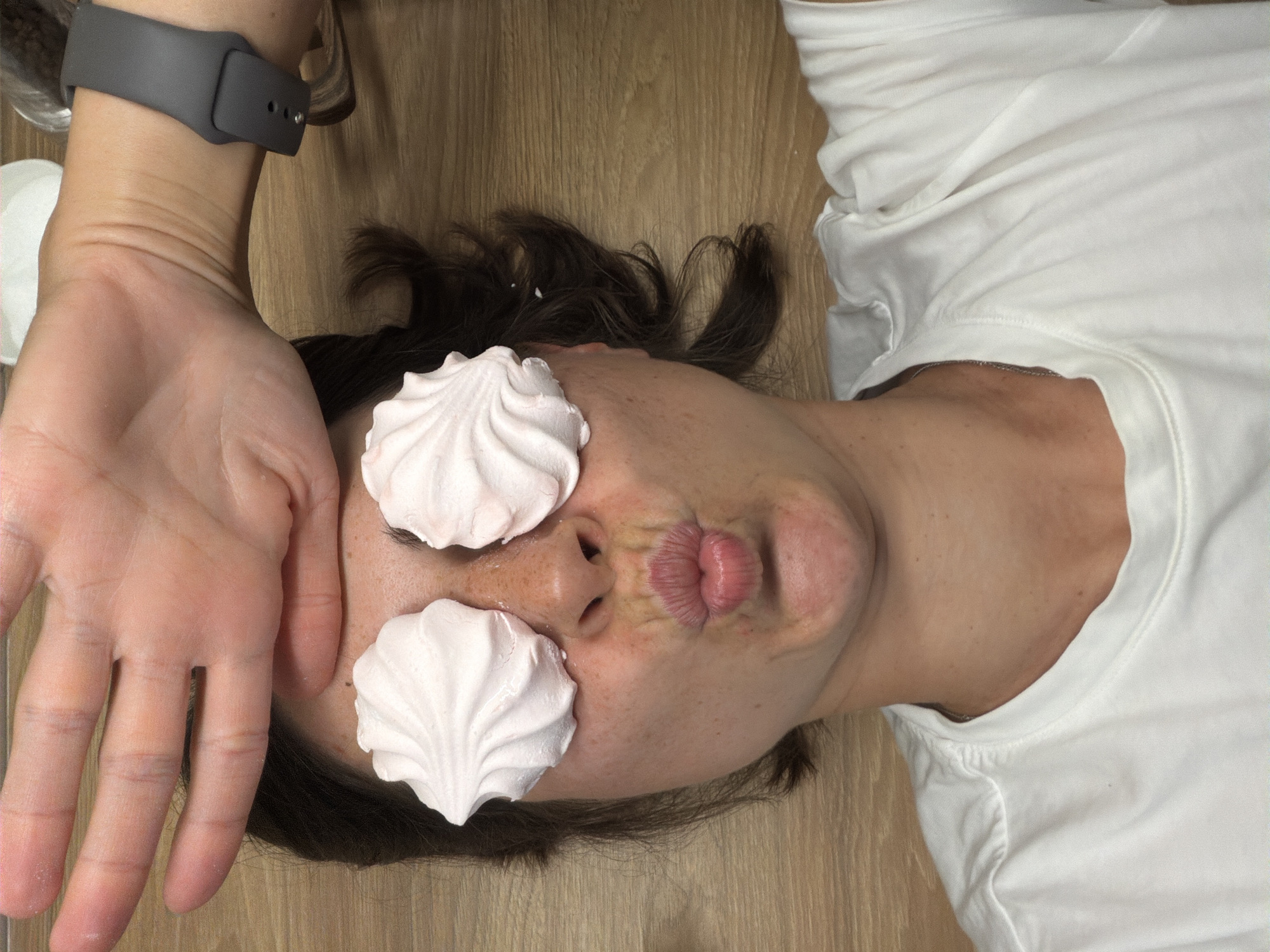 A woman is lying on a light brown, wood-grained surface wearing a white t-shirt. She has dark hair, and a gray smartwatch is visible on her left wrist. Two white, meringue-like objects are placed over her eyes. Her mouth is puckered. She is holding her left hand up to her forehead. A glass container is partially visible to the left of her arm. The location is not evident from this image.
