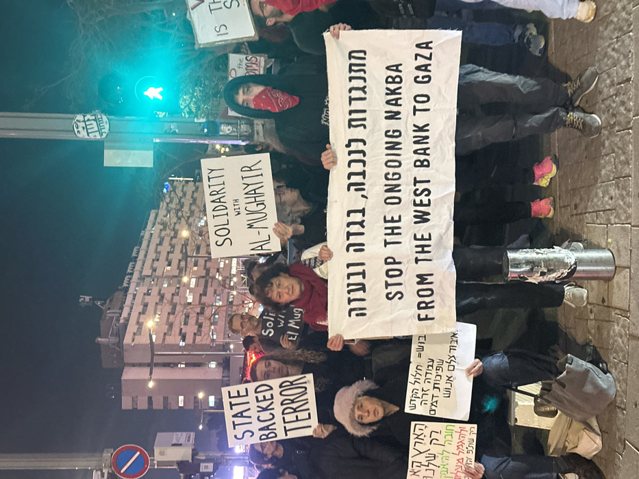 A nighttime street protest features individuals holding signs. One large banner states, "STOP THE ONGOING NAKBA FROM THE WEST BANK TO GAZA," also written in Hebrew. Other signs declare "SOLIDARITY WITH AL-MUGHAYIR," "STATE BACKED TERROR," and a few in Hebrew addressing loss of human dignity, desecration, and bloodshed. A masked person is present, and a traffic light glows green in the background.