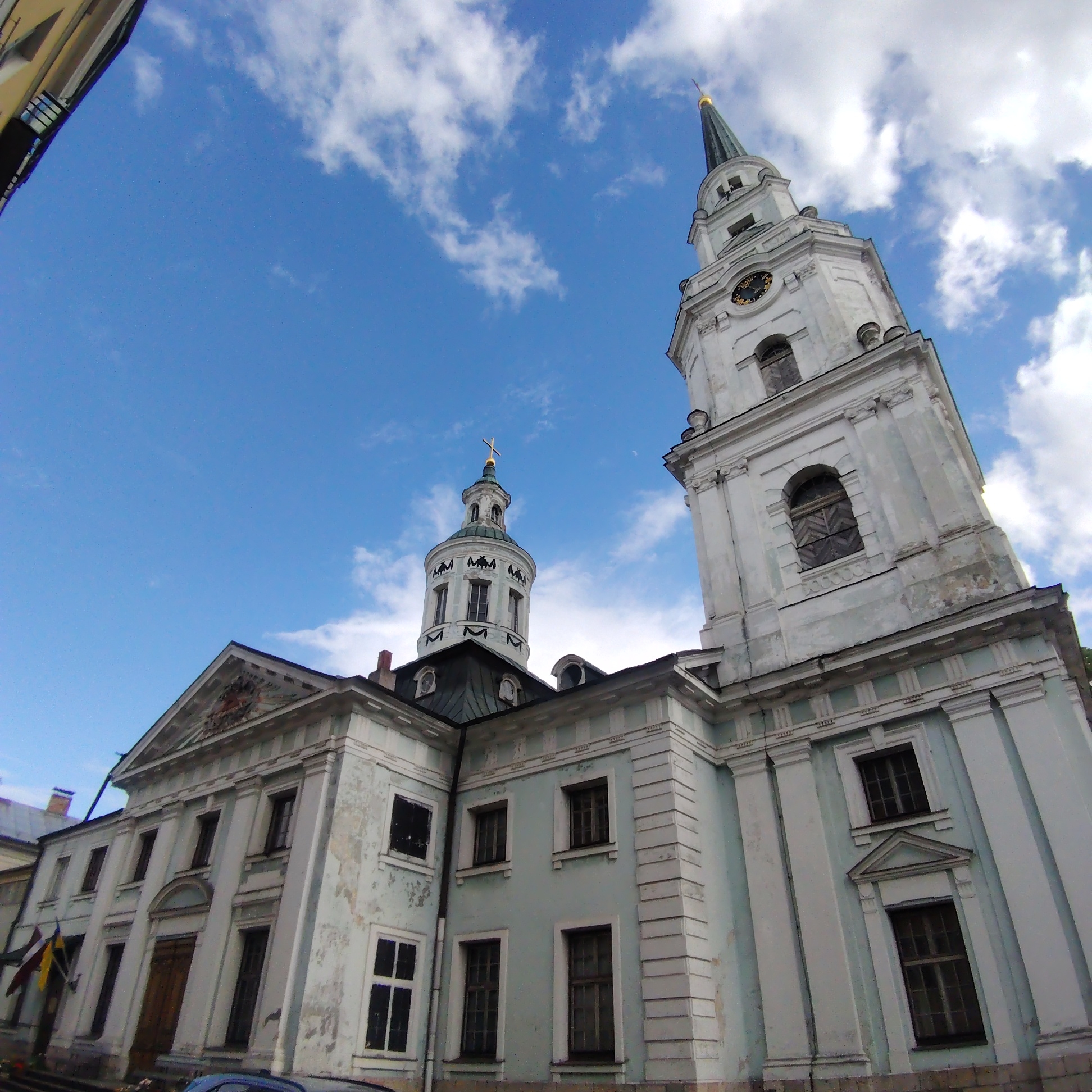 The image depicts the Church of St. Peter in Riga, Latvia, seen from a low angle. The architectural style is Baroque. The church comprises two towers, one taller than the other, both featuring clocks and spires. The main building exhibits classical architectural elements with a facade of light-colored stone or plaster, showing signs of age and weathering. The sky is blue with scattered clouds. A portion of an adjacent building can be seen on the left edge of the frame. A car is visible in the foreground. Flags are visible at the base of the church.