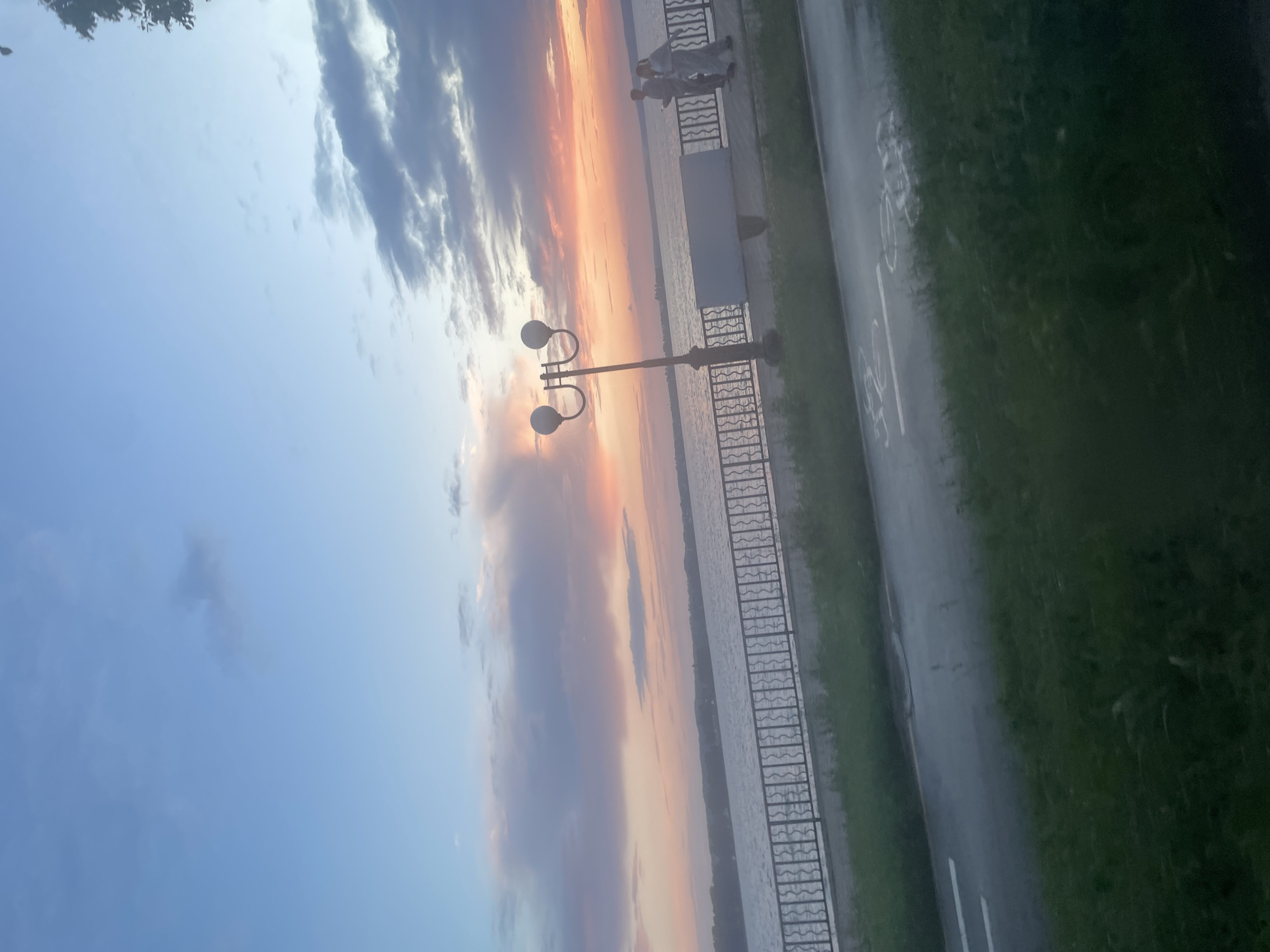 The image depicts a waterside promenade at dusk in Votkinsk, Russia. The sky is a gradient of pale blue to pink and orange hues, indicating sunset. Two individuals, a man and a woman, are walking side-by-side on the paved promenade. A decorative lamppost stands to their left. A metal railing separates the promenade from the calm body of water. Across the water, the distant shoreline is visible, lined with trees and some structures. In the foreground, a paved path, likely a bike lane indicated by faint white markings, runs parallel to the grass. The overall atmosphere is peaceful and serene.