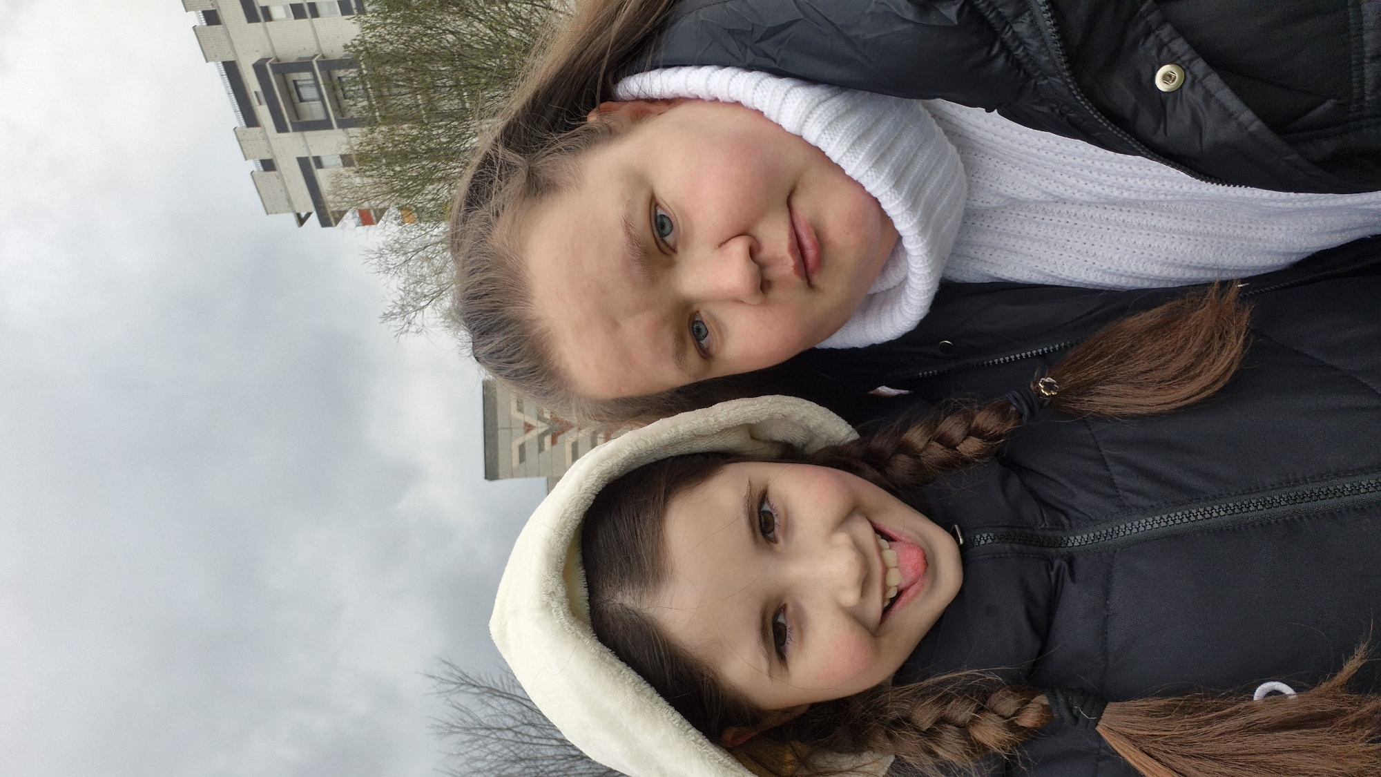 A close-up, vertically oriented outdoor portrait depicts two females, a child and an adult or older teen, against a gray, overcast sky and a distant building. The image is rotated 90 degrees counter-clockwise.
In the foreground, the younger girl smiles broadly, her tongue playfully sticking out slightly. She has brown eyes, rosy cheeks, and dark brown hair styled in two braided pigtails, visible against her black puffer jacket. She wears a cream or beige hooded jacket, possibly with a fleece lining, over the puffer jacket.
Behind and slightly above her is the older female, who looks directly at the camera with a neutral yet gentle expression, a faint smile playing on her lips. She has light blue eyes, rosy cheeks, and light brown hair. She is dressed in a white, ribbed turtleneck or scarf peeking out from under a black puffer jacket.
The background is largely taken up by the dull, cloudy sky, indicating an overcast day. To the upper right, part of a multi-story, modern apartment building with balconies is visible, along with bare tree branches, suggesting a cold season. This setting is located in Chaikovsky, Russia. The overall mood conveyed is one of warmth and affection between the two individuals despite the cold weather indicated by their attire and the bare trees. No text is visible in the image.