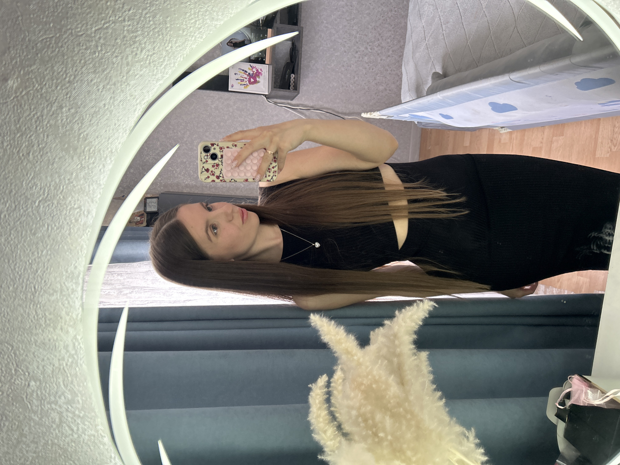 A woman stands in front of a mirror taking a selfie with her phone. The mirror has a decorative ring of light around it. The woman has long brown hair and is wearing a black dress with a cutout at the waist. Behind her, one can see furniture, including a shelf, a bed or ironing board, and some dried floral decorations. The location, Klin, Russia, cannot be verified from the image.