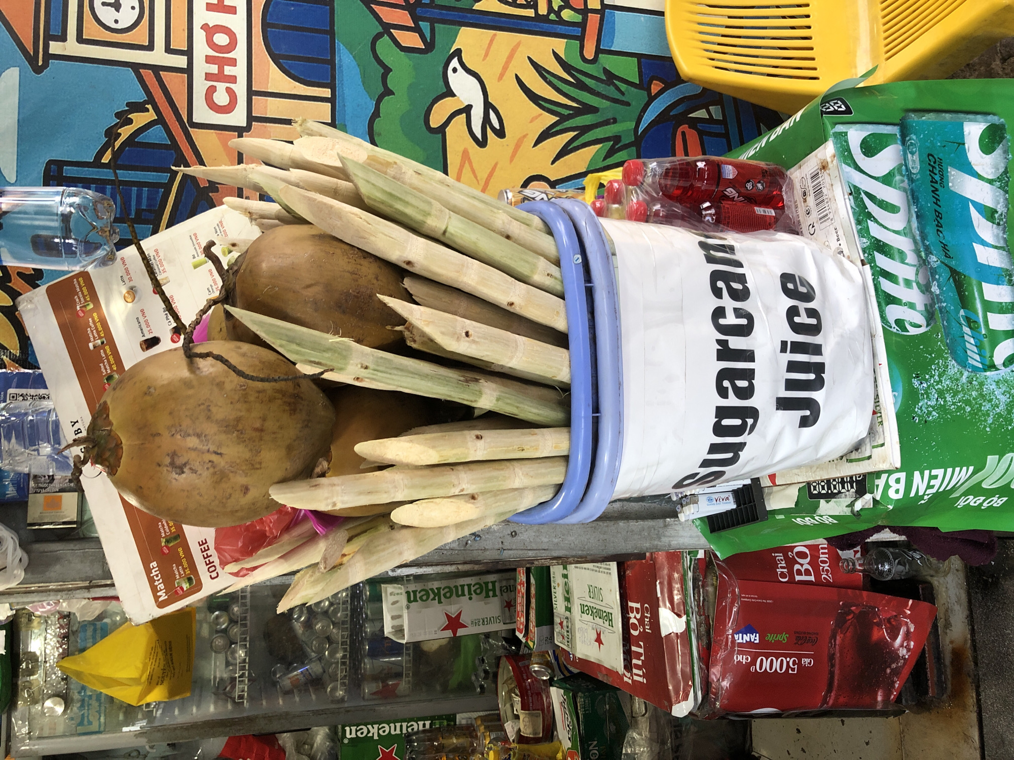 This image depicts a street-side vending stall, likely in the city of Đà Nẵng, Vietnam, specializing in fresh sugarcane juice. Dominating the foreground is a large white bucket with a blue rim, filled with freshly cut sugarcane stalks. The bucket has text printed on it, clearly visible as "Sugarcane Juice." Two coconuts are nestled amongst the sugarcane stalks, suggesting they might also be sold as beverages or an ingredient. Behind the bucket, a vibrant mural of a town with a clock tower and a white bird is visible. To the left of the stall, various products are displayed, including cans of Heineken, Coca-Cola, and Sprite, along with what appears to be a refrigerator unit. A bright yellow plastic chair is positioned to the right. The scene is cluttered, typical of a busy local market or roadside vendor, with a mixture of fresh produce and packaged drinks. The overall impression is one of a bustling commercial environment focused on refreshing drinks.
