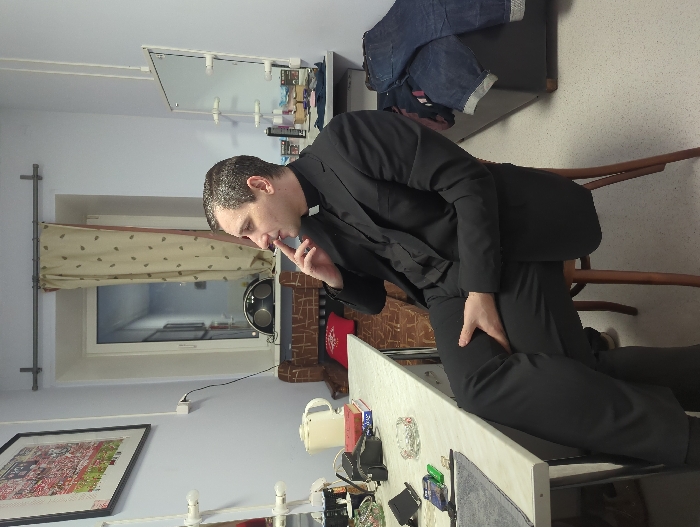 A male individual, appearing to be in his 30s to 40s, with short dark hair, is seated on a wooden stool within an interior room. He is dressed in a black clerical suit, comprising a black shirt with a white clerical collar, a black jacket, and black trousers. Positioned facing right in the frame, his head is turned slightly to the right. His left hand is raised, with his index finger near his mouth and thumb near his chin, a gesture consistent with holding a small item such as a cigarette. His right hand rests on his right thigh, and his legs are crossed.
The room features light blue walls and a light-colored, speckled floor. To the left, a light-colored desk holds a white electric kettle, stacked items, an empty glass ashtray, and a gray fabric. A framed picture depicting a crowded scene in red and white hangs above the desk. A window with a patterned beige curtain is visible in the background, offering a view into an interior hallway or another room. Below the window, a dark speaker system is present. To the right, a vanity mirror with exposed light bulbs is visible, with various toiletries on the surface beneath it. A dark grey sofa or bench, draped with blue denim jeans and other clothing, occupies the far right. The precise location within the city of Moscow, Russia, is not identifiable from the visual content of the image, but it depicts an indoor private or utility space.