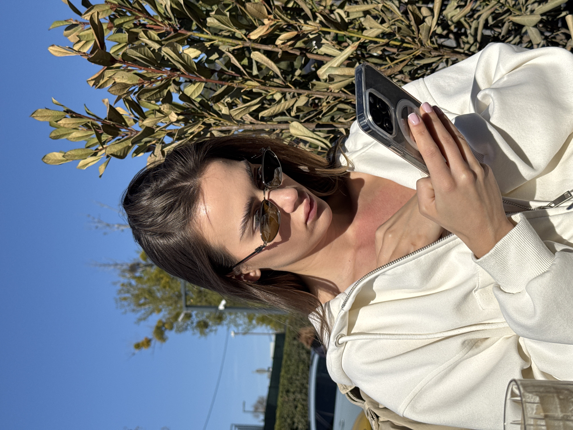 A young woman is captured in a close-up, outdoor shot on a bright, sunny day. She is positioned on the right side of the frame, looking downwards with a focused expression at a smartphone held in her right hand. She has long, dark hair and is wearing stylish sunglasses with dark, possibly reflective, lenses. Her attire consists of a light-colored, likely cream or white, zip-up hoodie, and her left hand is visible resting near her chest.
The smartphone features a clear protective case, and a circular MagSafe-like accessory is visible on its back. Her fingernails are neatly manicured with light-colored polish and French tips. In the upper left portion of the image, a dense mass of green and brown leaves from a tree or bush provides a natural backdrop against a clear, vibrant blue sky that dominates the upper and left parts of the frame. The background also shows blurred elements of an urban or suburban environment, including distant buildings and what appears to be a hedge or fence. A portion of a clear glass or cup is visible in the bottom right corner.
The strong, direct sunlight indicates it is daytime, likely mid-day, under clear weather conditions. The woman appears calm and engrossed in her phone activity. No visible text is discernible anywhere in the image. The scene suggests a relaxed outdoor setting, possibly a cafe or park.