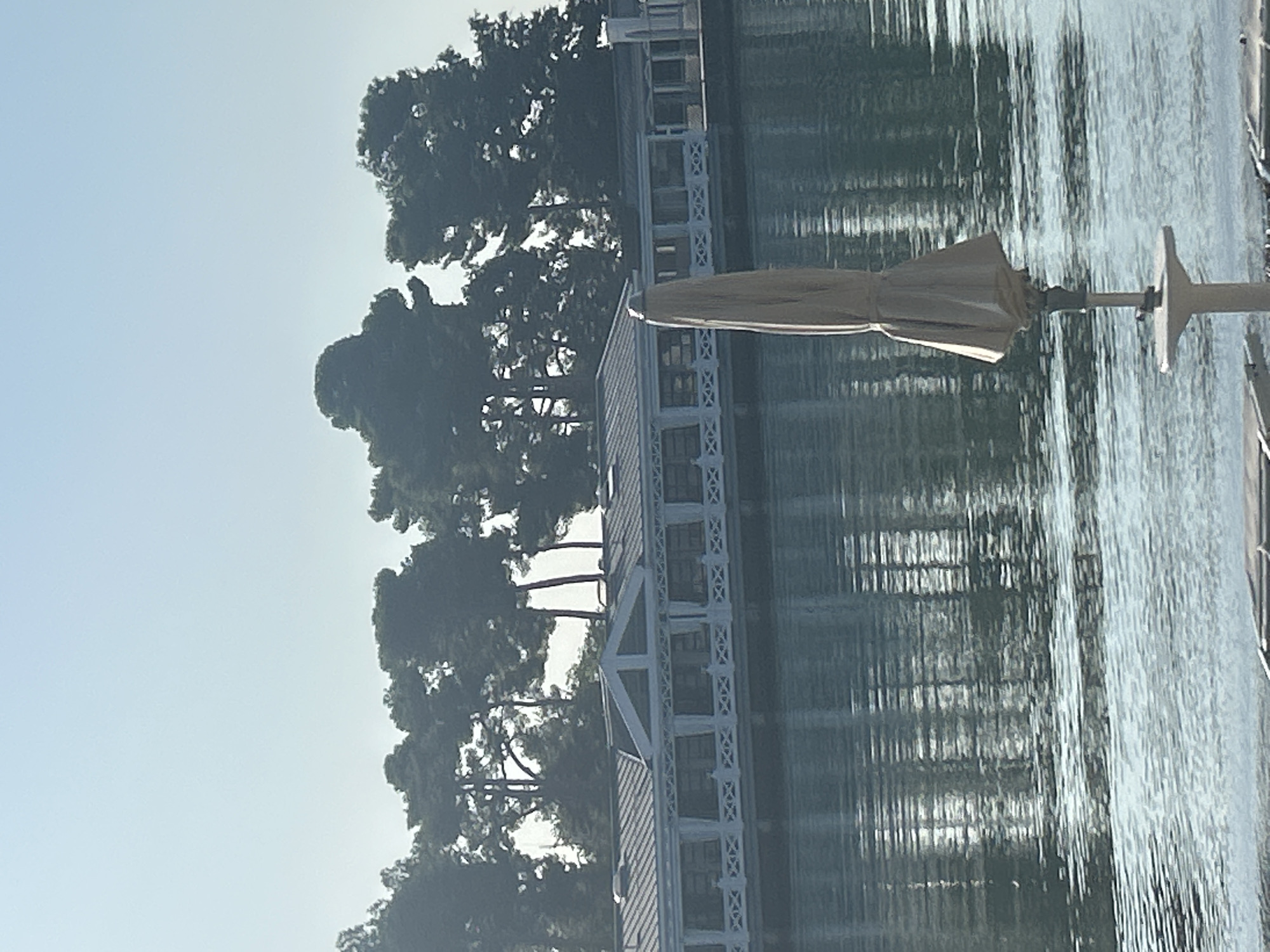 A closed, light-tan patio umbrella stands on a small circular base with a pedestal support in the foreground. Behind it, a long, white, low-rise building with a dark roof is situated on the water's edge. The building has a series of large windows and a decorative railing. A line of dark green trees with thin trunks forms a backdrop behind the building. The water in front of the building reflects the building and the trees, creating a shimmering effect. The sky is a clear, pale blue. This scene is likely located at the Hotel Adriatic in Umag, Croatia, which is known for its waterfront location and buildings that match this description.
