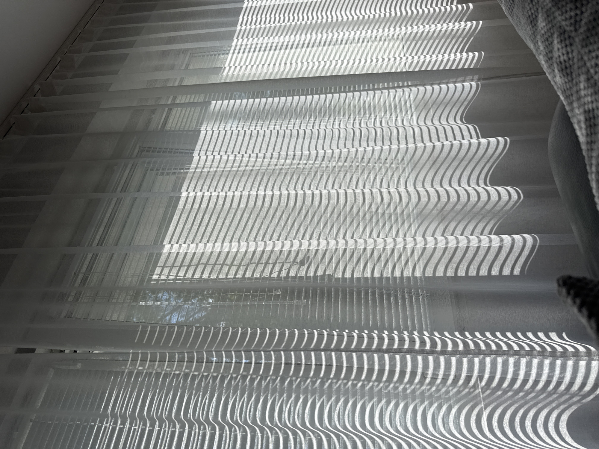The image shows light filtering through horizontal blinds or shutters, creating a pattern of alternating light and shadow. The light appears to be strong sunlight, casting distinct shadows with curved lines. The background visible through the blinds seems to show outdoor scenery. The presence of blinds suggests an indoor environment, likely a window. There is a couch on the right side of the image. The image was taken in Lovech, Bulgaria.