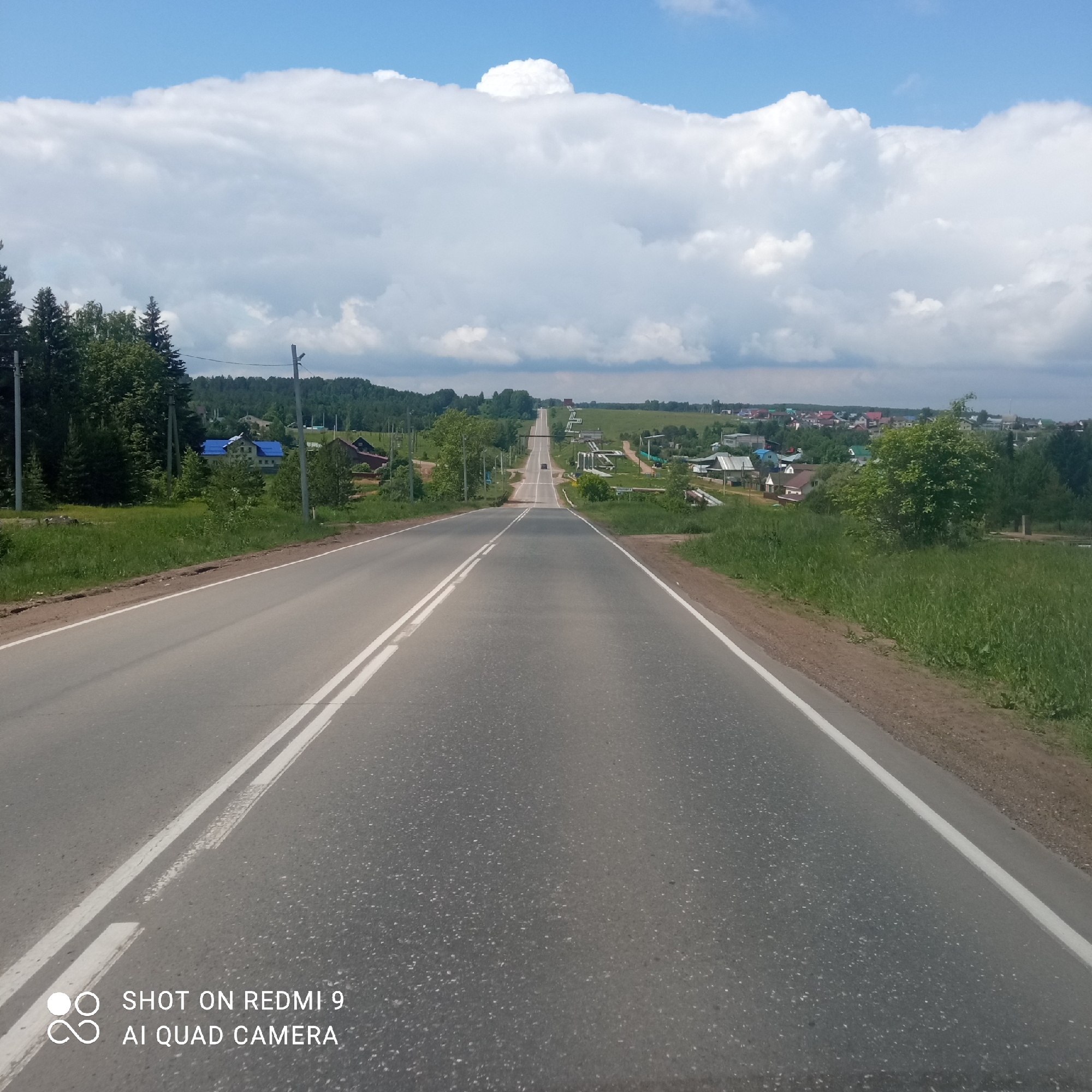 The image presents a wide-angle view of a rural landscape, likely in Ольховка, Russia, dominated by a long, asphalt road stretching into the distance. The road, marked with double white lines in the center and a single white line on the right shoulder, appears to undulate over gentle hills, creating a sense of depth and perspective. It is empty of traffic, suggesting a quiet moment on a country route.
To the left of the road, a dense treeline of evergreen and deciduous trees forms a green border, with several utility poles running alongside. Beyond the trees, glimpses of houses with colorful roofs, including a distinct blue-roofed building, are visible, indicating a village or settlement. On the right side of the road, an expanse of green grass and scattered bushes slopes gently upwards, with more residential structures and utility lines discernible in the mid-ground and background. The distant horizon reveals a sprawling village nestled amidst the greenery.
The sky is a mix of bright blue patches and large, white cumulus clouds, casting soft shadows but indicating fair weather conditions. The overall impression is one of a bright, sunny day, possibly late spring or summer, in a serene countryside setting.
In the bottom left corner of the image, white text overlays the road, stating: "SHOT ON REDMI 9 AI QUAD CAMERA".