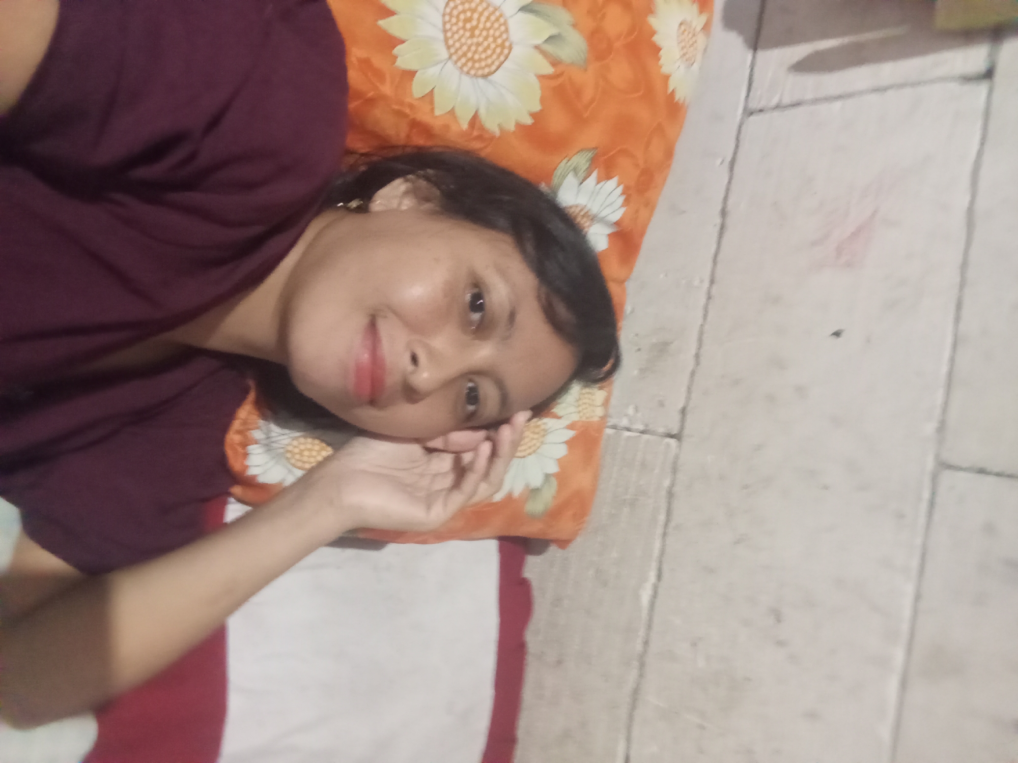A young woman is depicted lying down indoors, resting her head on a vibrant floral pillow. She is positioned diagonally across the frame, looking directly at the camera with a gentle, relaxed smile. She has dark hair, dark eyes, and a hint of lipstick on her lips. She is wearing a dark maroon short-sleeved top. Her left hand is casually placed near her face, possibly supporting her head.
The pillow features a bright orange background adorned with prominent white and yellow sunflower-like patterns. Below the woman, a portion of a white and red striped blanket or sheet is visible. To her right, a light-colored tiled floor with visible grout lines occupies a significant part of the background, suggesting a typical indoor setting. The ambient lighting indicates an indoor scene, likely in a home or similar dwelling. The overall impression is one of calm and relaxation, captured in Baron, Indonesia. There is no visible text in the image.