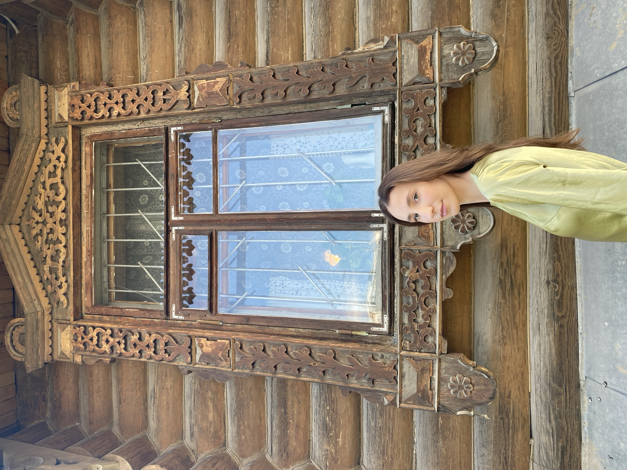 A young woman is standing next to a window with ornate wooden carvings at the Vysotsky Business Center in Yekaterinburg, Russia. The window is set in a wall constructed from logs. The girl wears a pale green top. The window has metal bars and reflections of the surrounding environment. The window frame is richly decorated with carved geometric and floral patterns. The exterior wall is made up of interlocking logs forming a textured surface. The area to the far right of the image is a gray, stone-like surface.