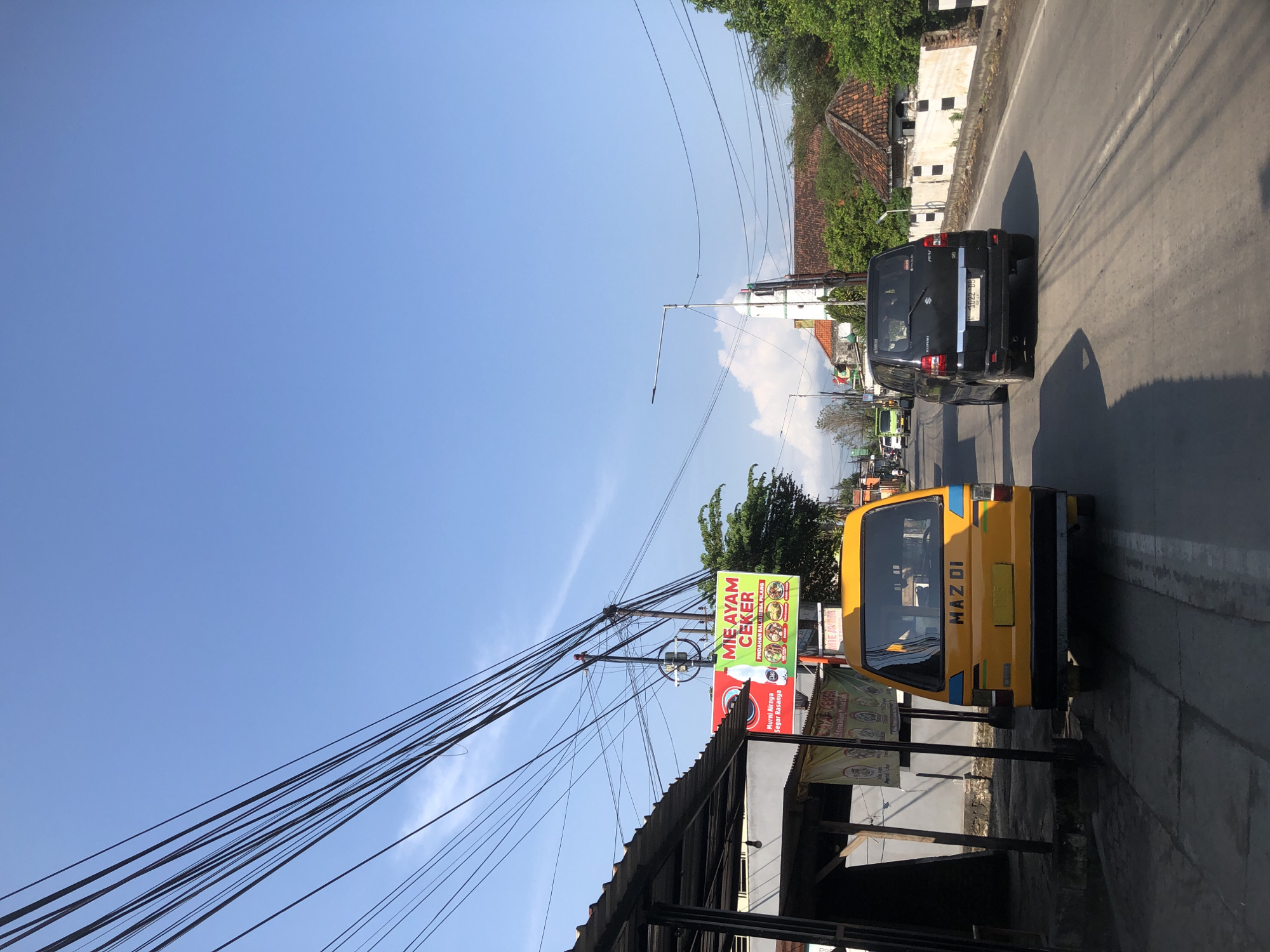 The image depicts a street scene in Modong, Indonesia, on a bright, sunny day under a clear blue sky. In the foreground, a yellow van with "MAZ DI" written on its rear is parked on the side of the road. To its left, a covered structure with metal supports is visible, with banners hanging from it. Across the street, a black SUV is driving away from the camera. Further down the road, a green truck is also traveling. A prominent green and red billboard advertising "MIE AYAM CEKER" (chicken noodle soup) is positioned near the yellow van. Power lines crisscross the sky, supported by utility poles. Buildings with terracotta roofs and whitewashed walls line the right side of the road, interspersed with lush green trees. In the distance, a white building with a green dome, possibly a mosque or church, can be seen. The road surface shows tire marks and shadows cast by the sun.