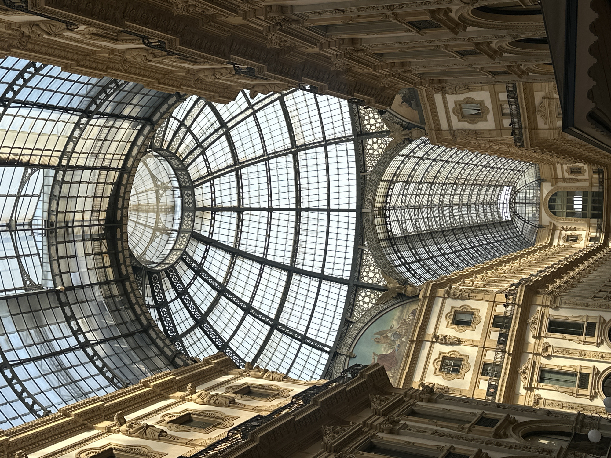 This is a low-angle, vertical shot of the Galleria Vittorio Emanuele II in Milan, Italy. The shot showcases the magnificent glass and iron dome of the Galleria, along with the ornate architecture of the surrounding buildings. The sunlight streams through the glass panels of the dome, illuminating the interior. The buildings on either side feature elaborate facades with decorative elements, balconies, and arched windows. A fresco painting is visible on one of the upper walls. There are no people clearly visible in the frame. The scene depicts the grandeur and architectural splendor of this historic Milanese landmark.