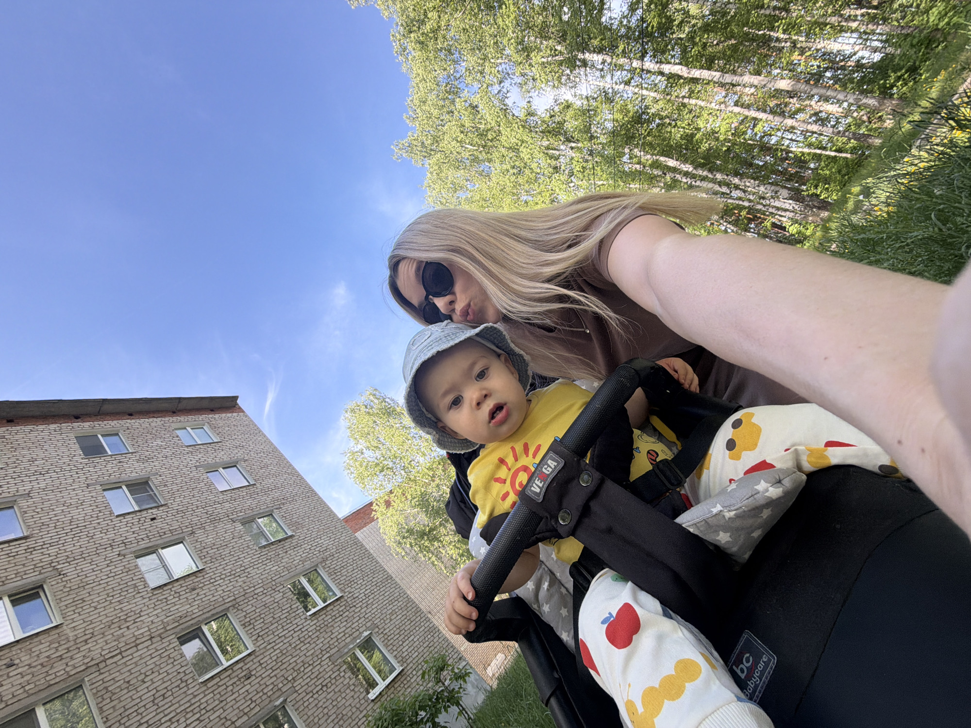 A woman with long blonde hair and sunglasses is taking a selfie with a baby in a stroller outdoors. The baby wears a sun hat and a yellow shirt with a sun graphic. The stroller is black and has patterned fabric inside. The background includes a light brick building with windows, trees with green leaves, and a blue sky with clouds. The location is likely Chaikovsky, Russia.