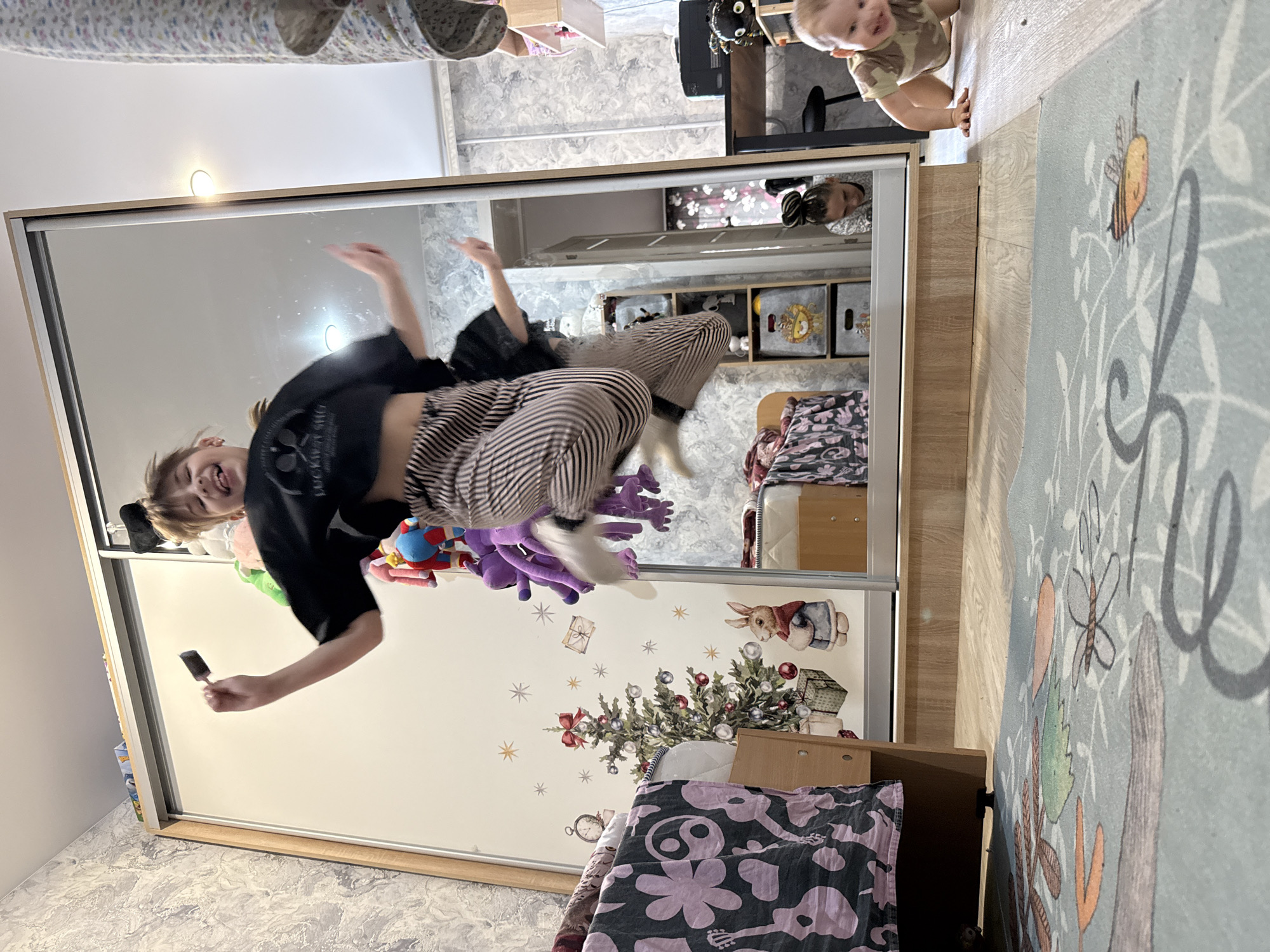 The image shows a mirrored wardrobe in a room. A young woman is jumping in front of the wardrobe with an object in her hand. The reflection shows a cluttered room with a child visible at the top of the mirror, and toys and decorations on the wardrobe surface. The exact location is unclear, but the room appears to be a child's bedroom.