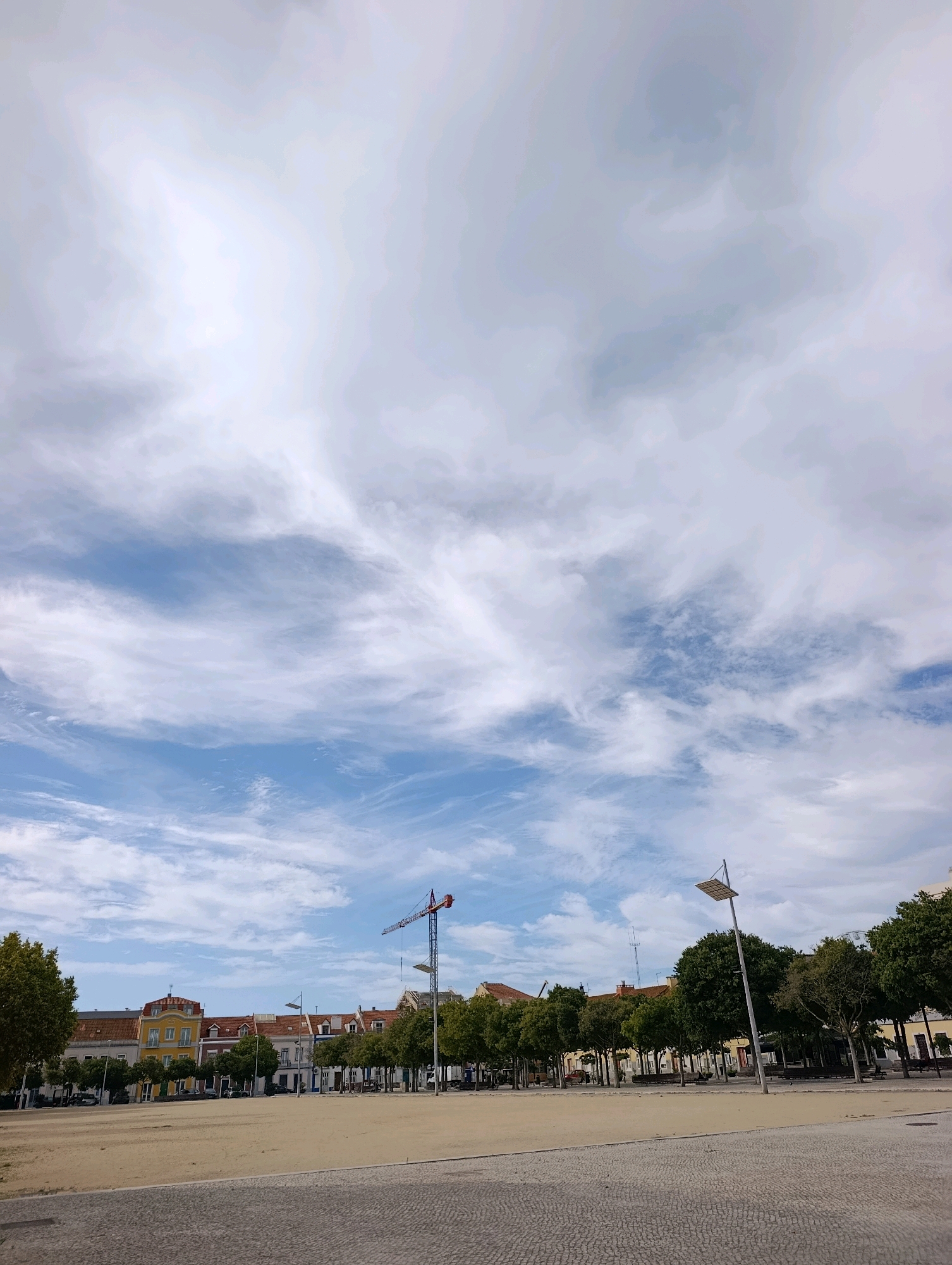 This is a wide-angle, eye-level, full shot of a public square in Setúbal, Portugal, on a partly cloudy day. The square is mostly empty, with a sandy ground in the foreground and a paved area in the middle distance. A row of trees lines the perimeter of the square, with buildings visible behind them. In the background, a construction crane stands tall against the sky, suggesting ongoing development in the city. The sky is filled with scattered clouds, some white and wispy, others darker and more textured, creating a dynamic atmospheric effect. The architecture of the buildings suggests a Mediterranean influence, with terracotta roofs and colorful facades.