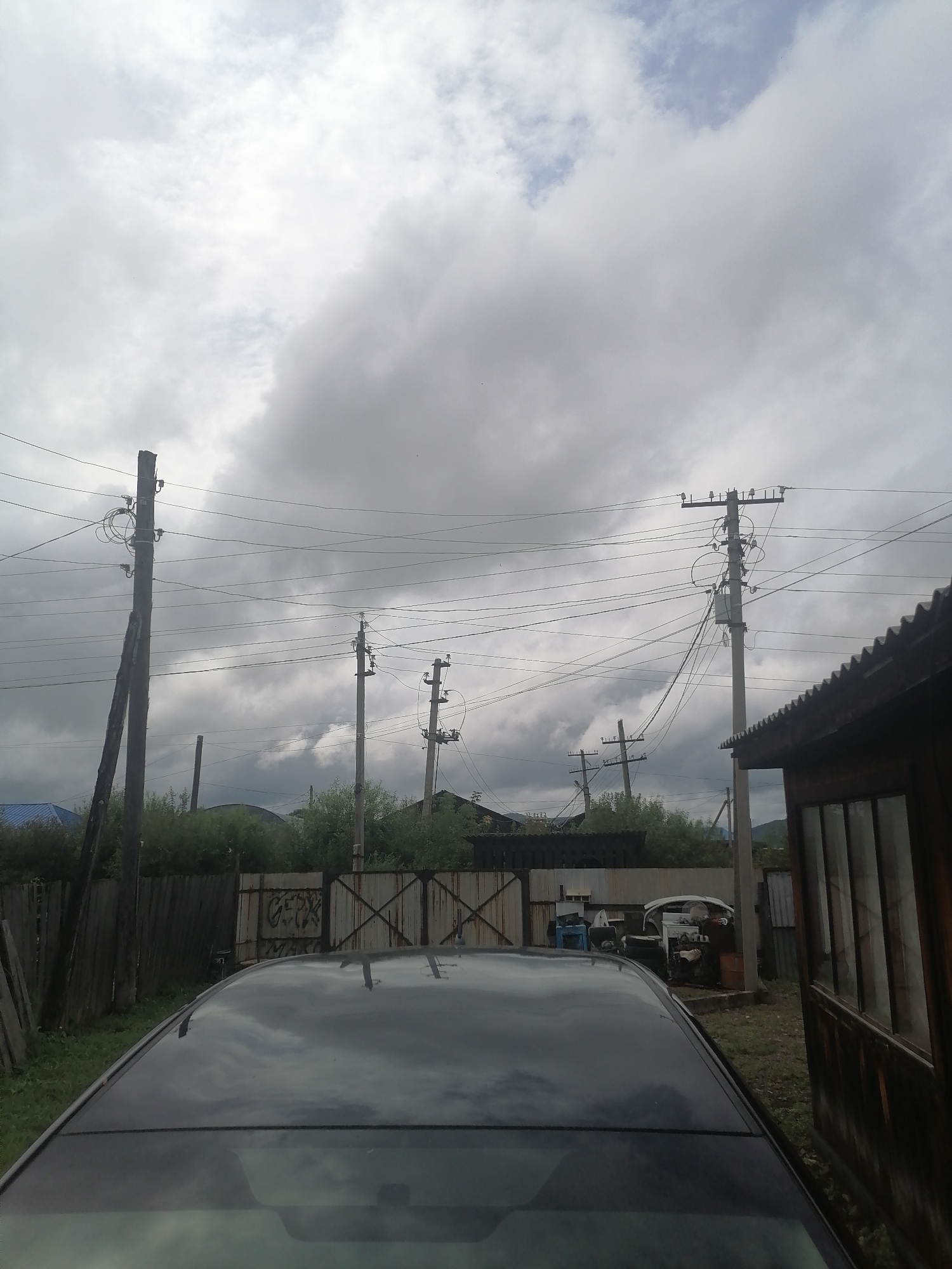 This image depicts a cloudy outdoor scene in what appears to be a residential or semi-rural area, possibly Novopokrovka, Russia, given the context. The foreground is dominated by the glossy black roof and windshield of a car, reflecting the overcast sky and surrounding elements.
In the mid-ground, a wooden fence runs horizontally across the frame, separating the car's location from what lies beyond. To the left of the fence, a tall, weathered wooden utility pole stands at an angle, supporting numerous electrical wires that stretch across the image. Behind this pole, a blue roof of a building is partially visible.
Further into the mid-ground, a series of concrete and wooden utility poles with electrical equipment are positioned, suggesting an electrical infrastructure network. Between these poles and behind the fence, there is dense green foliage and trees.
In the right mid-ground, a rustic wooden structure with a corrugated roof is present, and to its left, a metal gate with visible graffiti is apparent. Beyond the gate, a collection of miscellaneous items are stored, including what looks like car parts or scrap metal, a blue plastic container, and a white structure that could be part of a vehicle or machinery.
The background is filled with a dramatic, heavily clouded sky, with patches of lighter sky peeking through. The overall atmosphere is subdued, consistent with overcast weather. No people are visible in the image. The scene appears to be an everyday, possibly slightly disheveled, domestic or utility area.