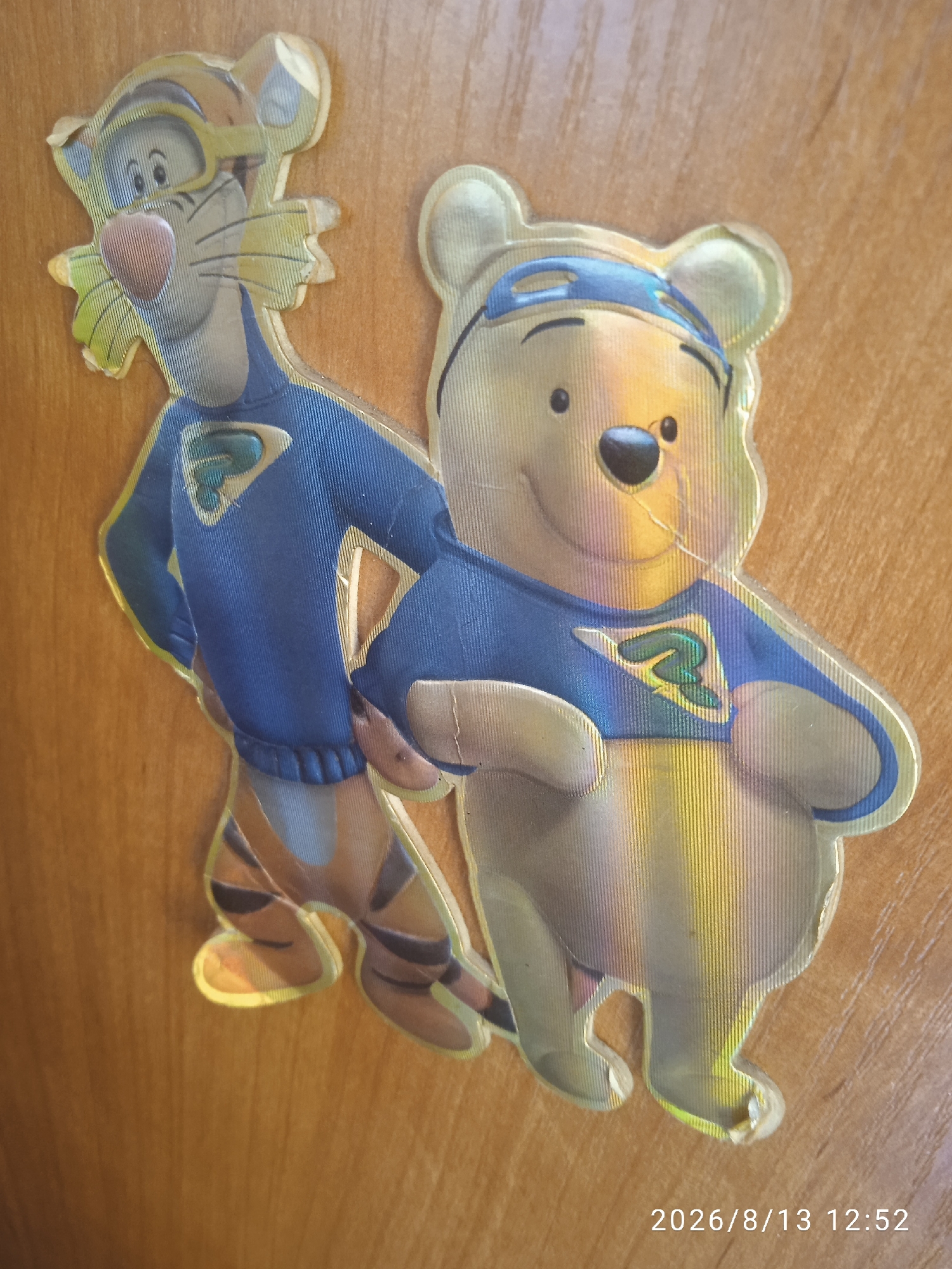 The image displays two holographic stickers of cartoon characters, Tigger and Winnie the Pooh, superimposed on a wooden surface. Tigger, on the left, is depicted wearing a blue outfit with a yellow emblem and yellow goggles. Winnie the Pooh, on the right, is also wearing a blue outfit with a similar emblem and a blue headband. Both characters are standing and appear to be in a dynamic pose. The stickers have a reflective, iridescent quality, causing light to refract and create rainbow-like effects. The wooden surface appears to be a table or a cabinet. In the bottom right corner of the image, a timestamp reads "2026/8/13 12:52". The image does not provide any information about a specific location in Chișinău, Moldova.