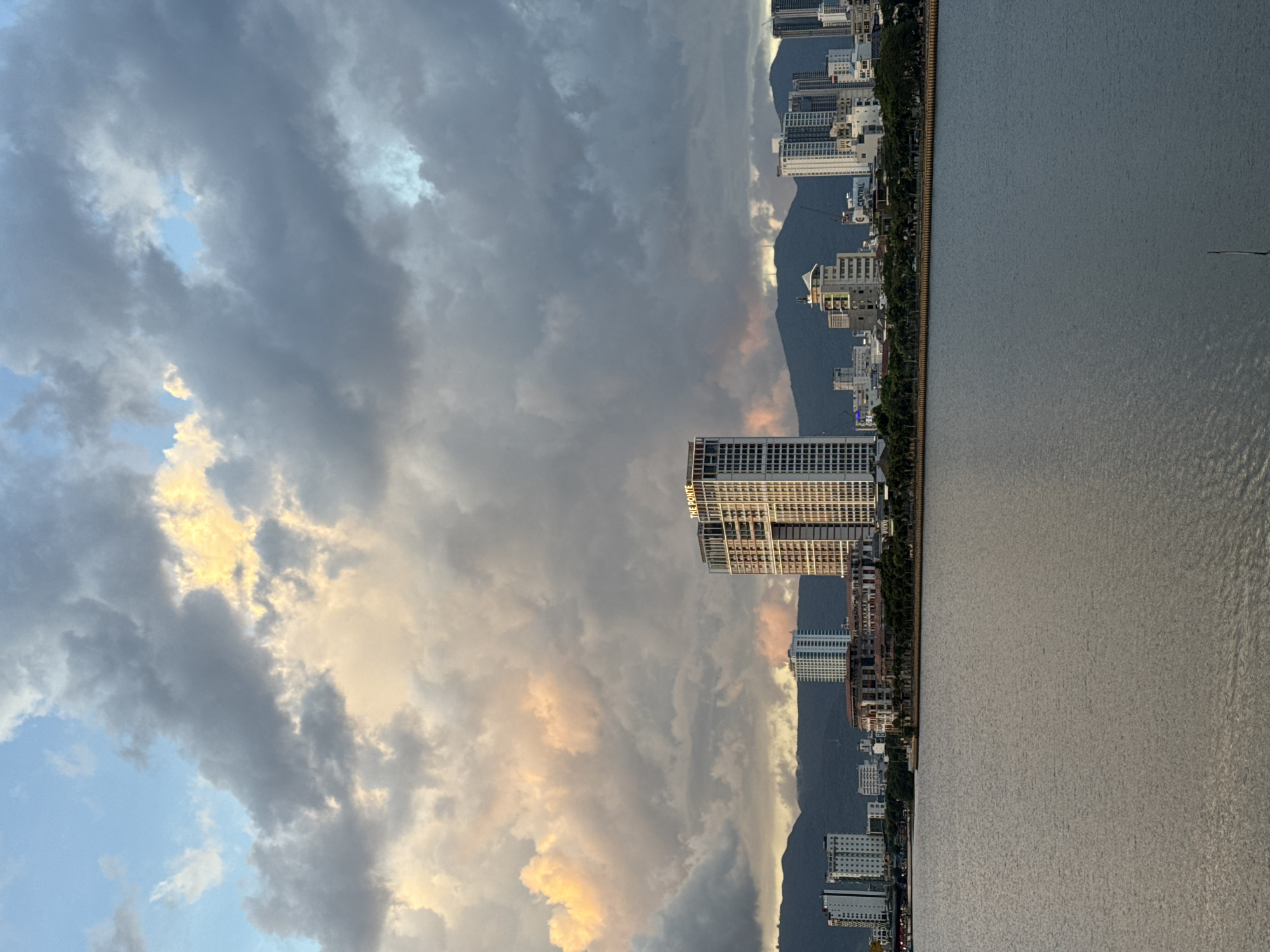 This is a landscape photograph of the Han River in Da Nang, Vietnam, taken at sunset. The sky is filled with dramatic clouds, tinged with pink and orange from the setting sun. In the distance, a city skyline rises, featuring numerous modern buildings of varying heights. A prominent building in the center of the frame displays the text "THE PONTE" on its upper facade. Beyond the city, a mountain range forms a dark silhouette against the colorful sky. The foreground is dominated by the tranquil surface of the Han River, reflecting the muted tones of the sky and cityscape. The overall scene suggests a peaceful urban evening.